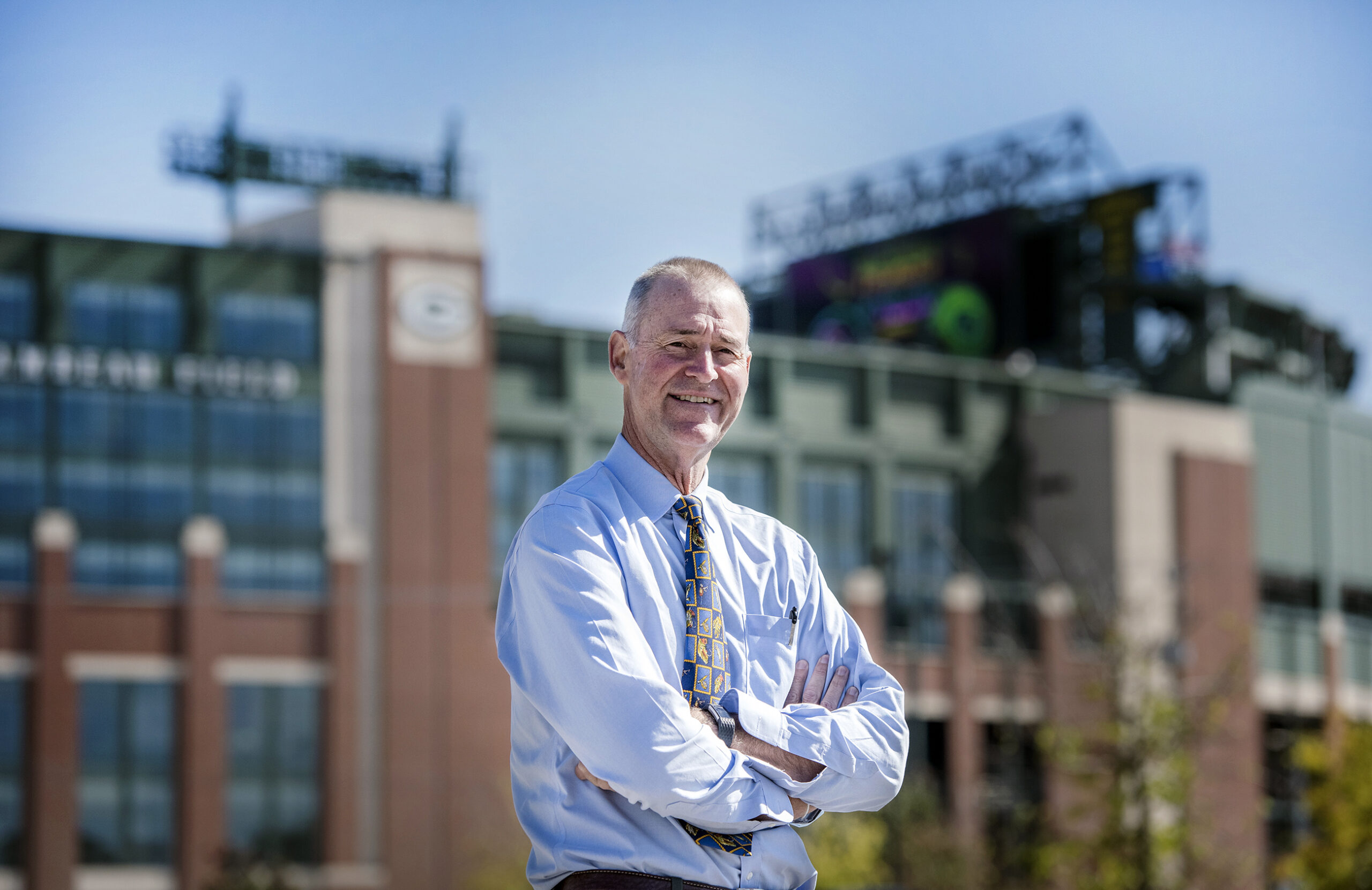 Dr. Anderson stands with his arms crossed on a sunny day. Lambeau Field can be seen behind him.