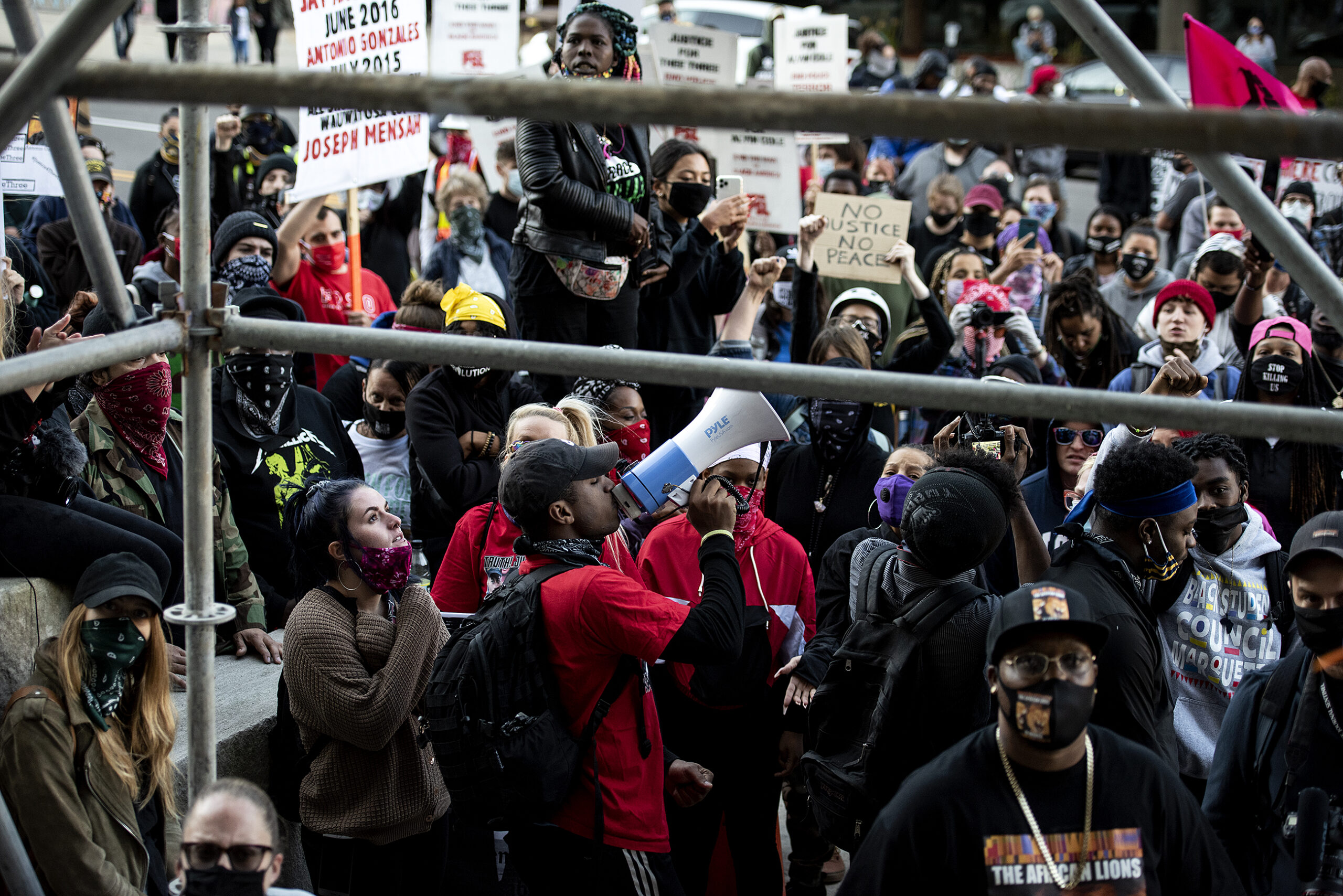 a man with a blue and white megaphone leads a sea of protesters in chants