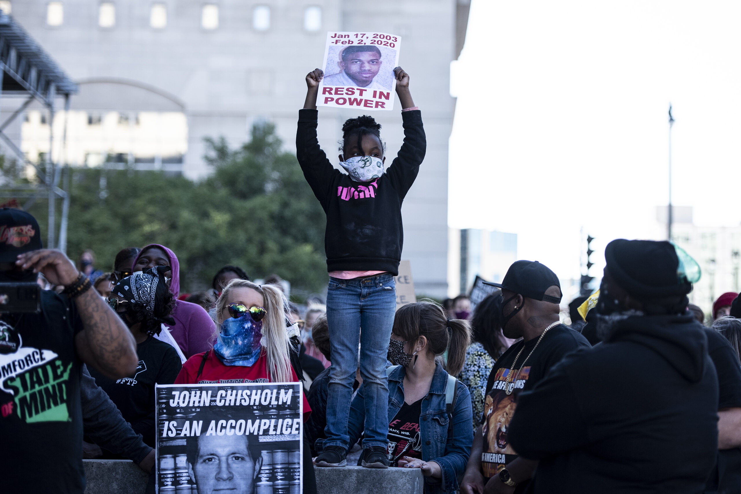 a child stands on a ledge above the crowd and holds a sign above her head