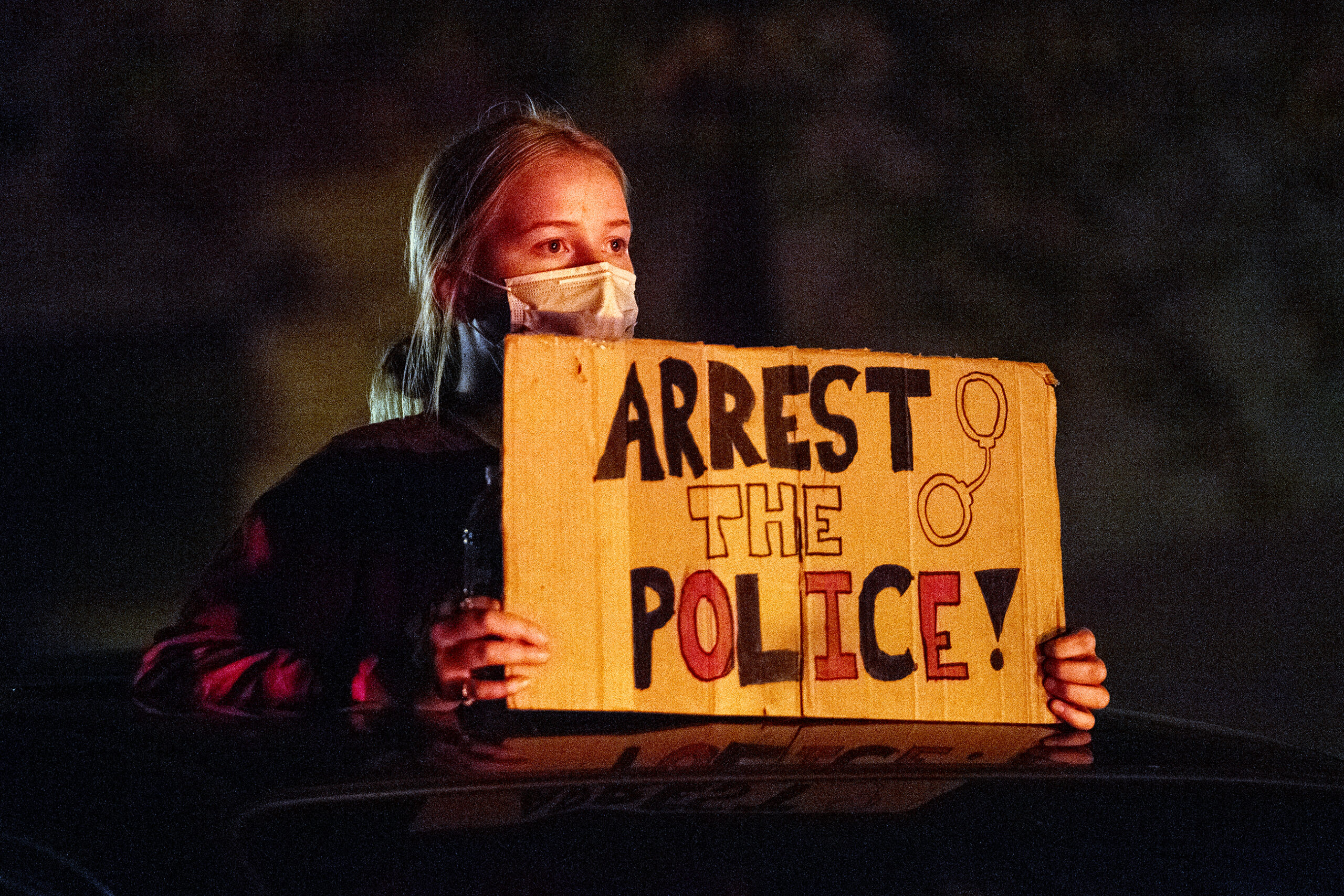 A protester holds a sign that says "arrest the police." She's is lit by a red tail light in an otherwise dark night.