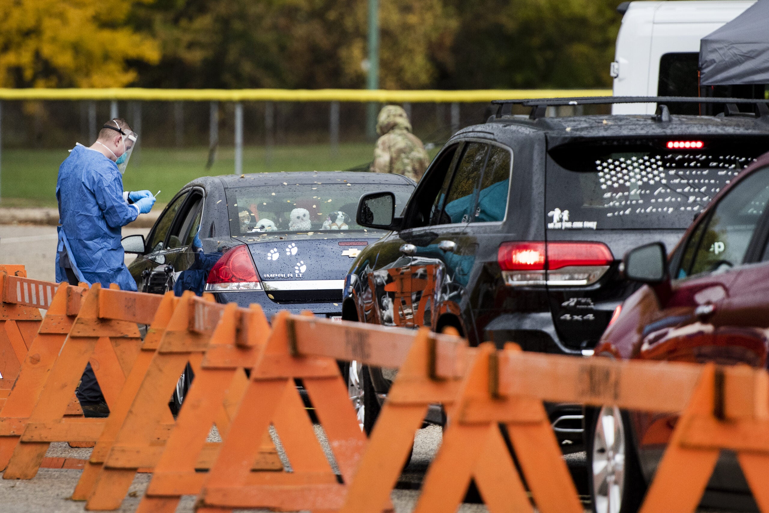 A line of cars next to orange barricades. A man in a blue gown and face shield stands next to one of them.
