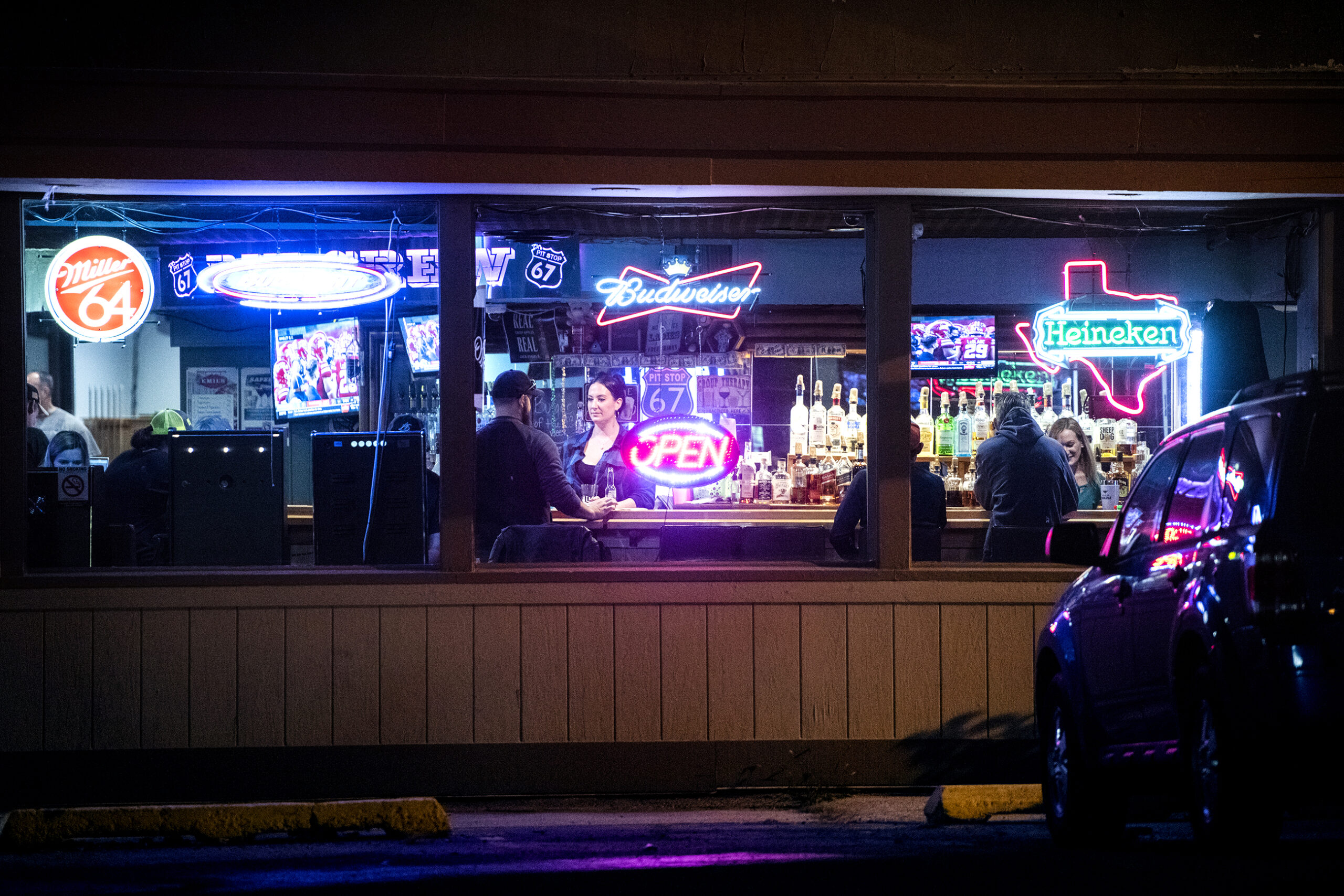 Neon signs illuminate the windows of a bar. a bar tender serving patrons can be seen inside.