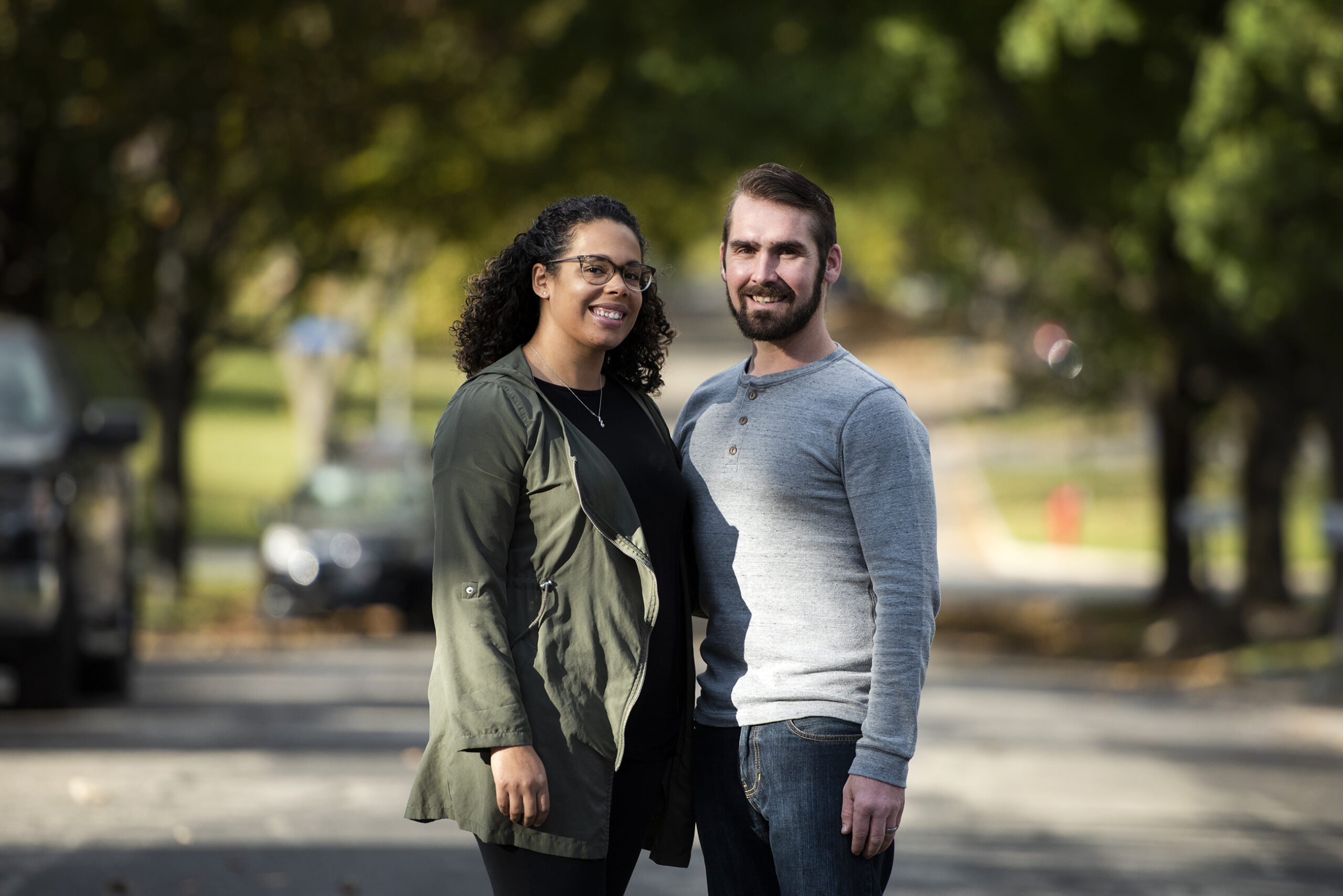 A couple stands together outside in the sunlight surrounded by green trees