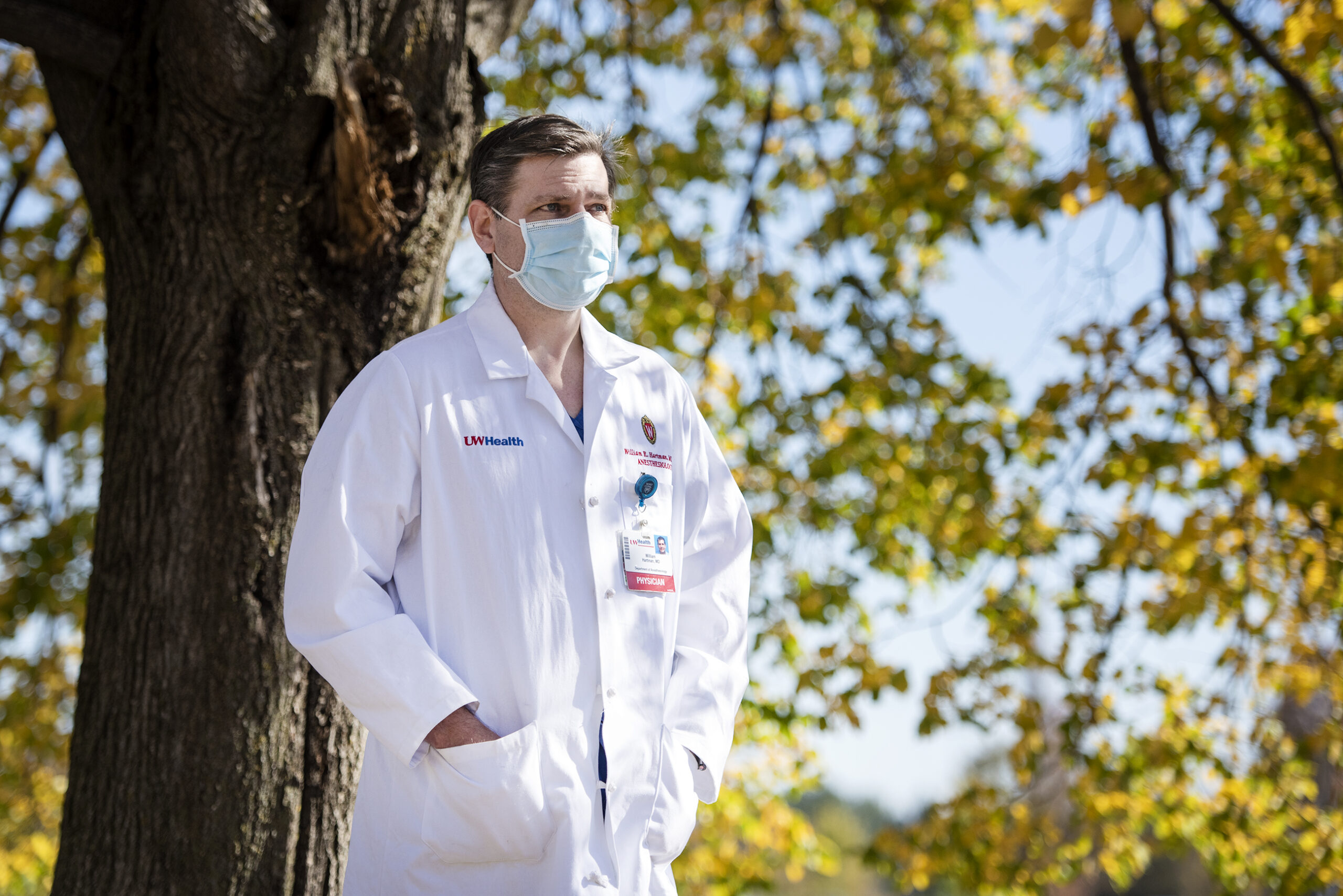 A doctor in a white coat and face mask stands in front of a tree with yellow fall leaves