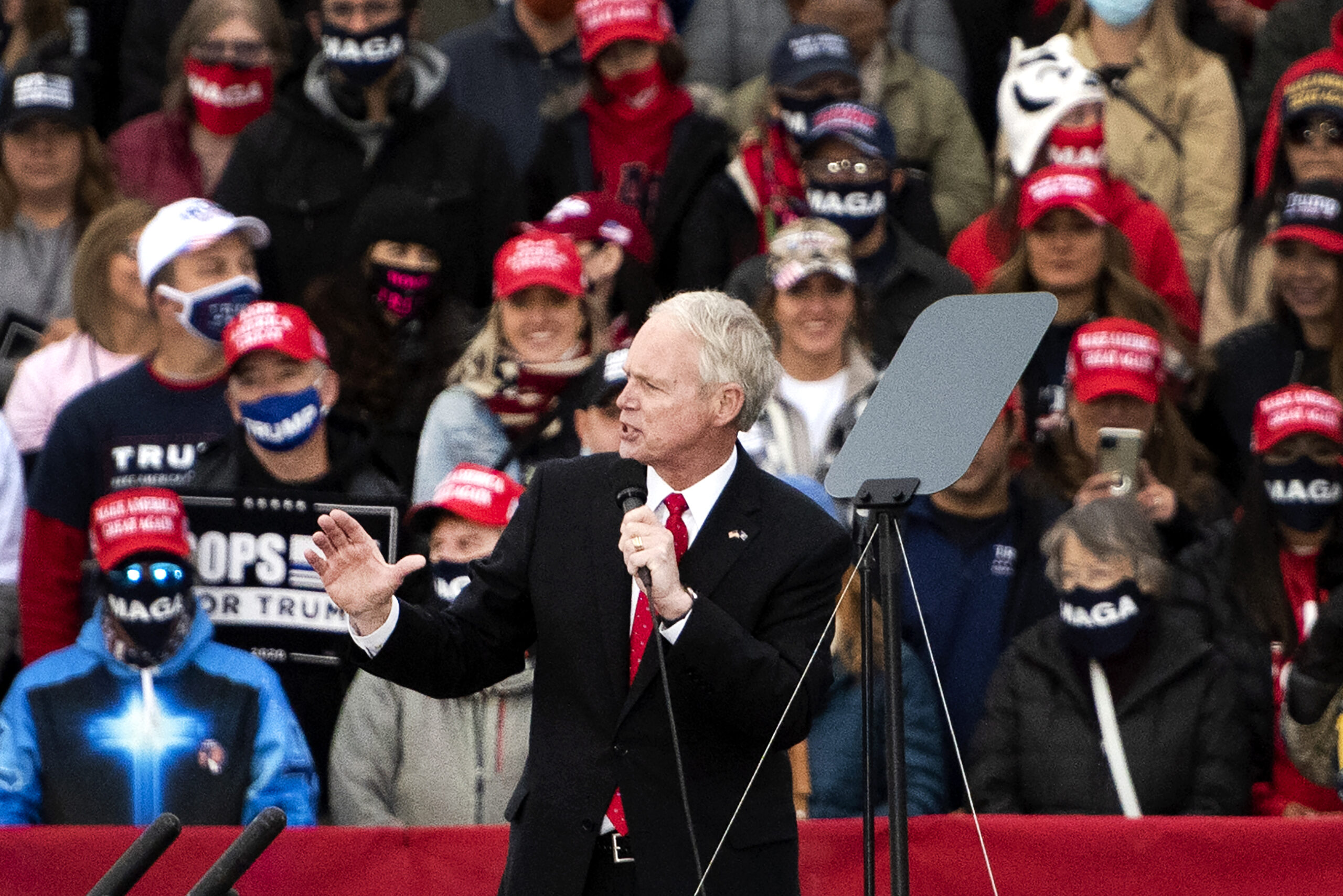 Trump supporters in MAGA hats can be seen behind Sen. Ron Johnson as he speaks into a microphone