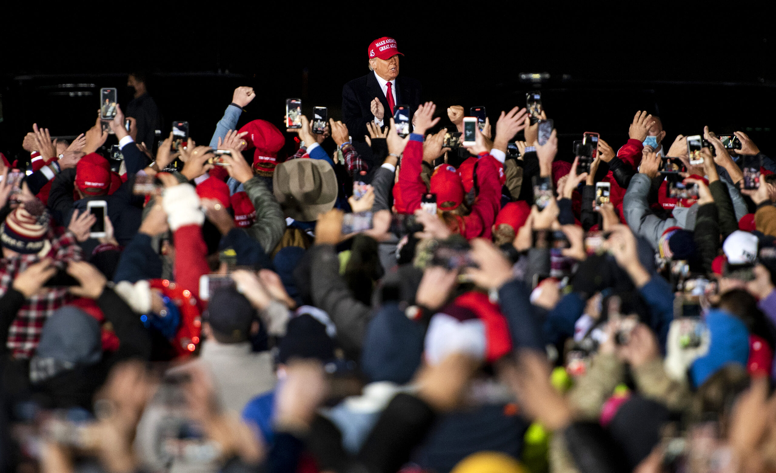 President Donald Trump wears a red hat as he approaches supporters who are gathered close together and raising their hands