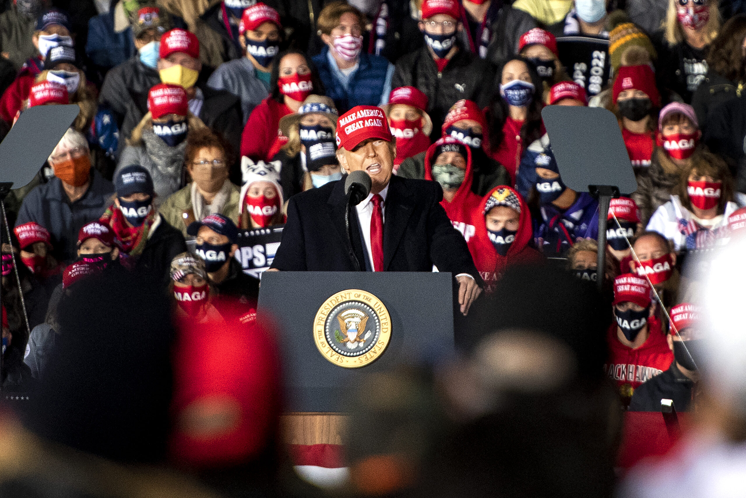 President Donald Trump stands on stage wearing a red MAGA hat as he speaks to supporters