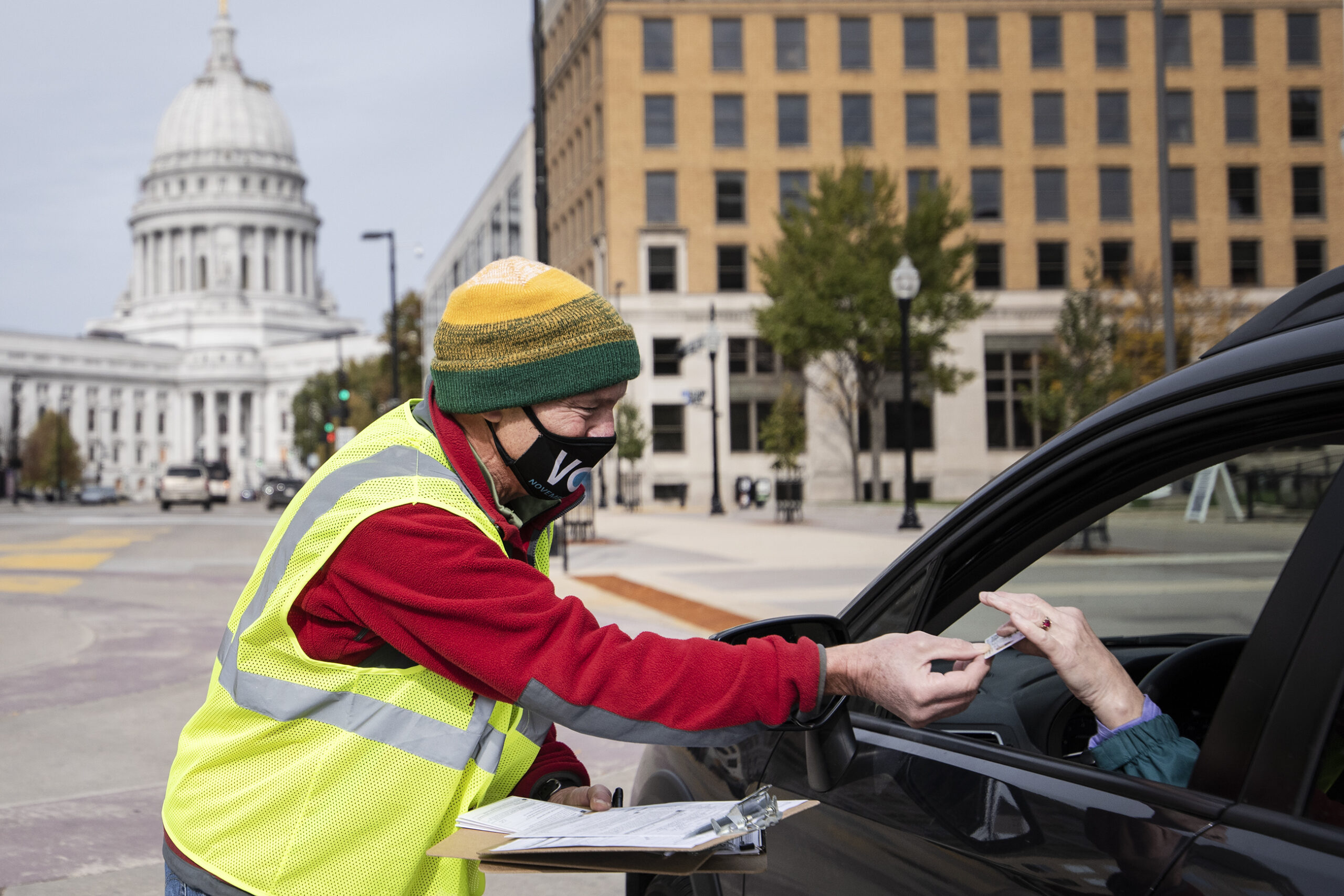 The Wisconsin State Capitol can be seen behind a poll worker in a mask and neon vest. He takes an ID card from a voter's hand as they sit in their car.