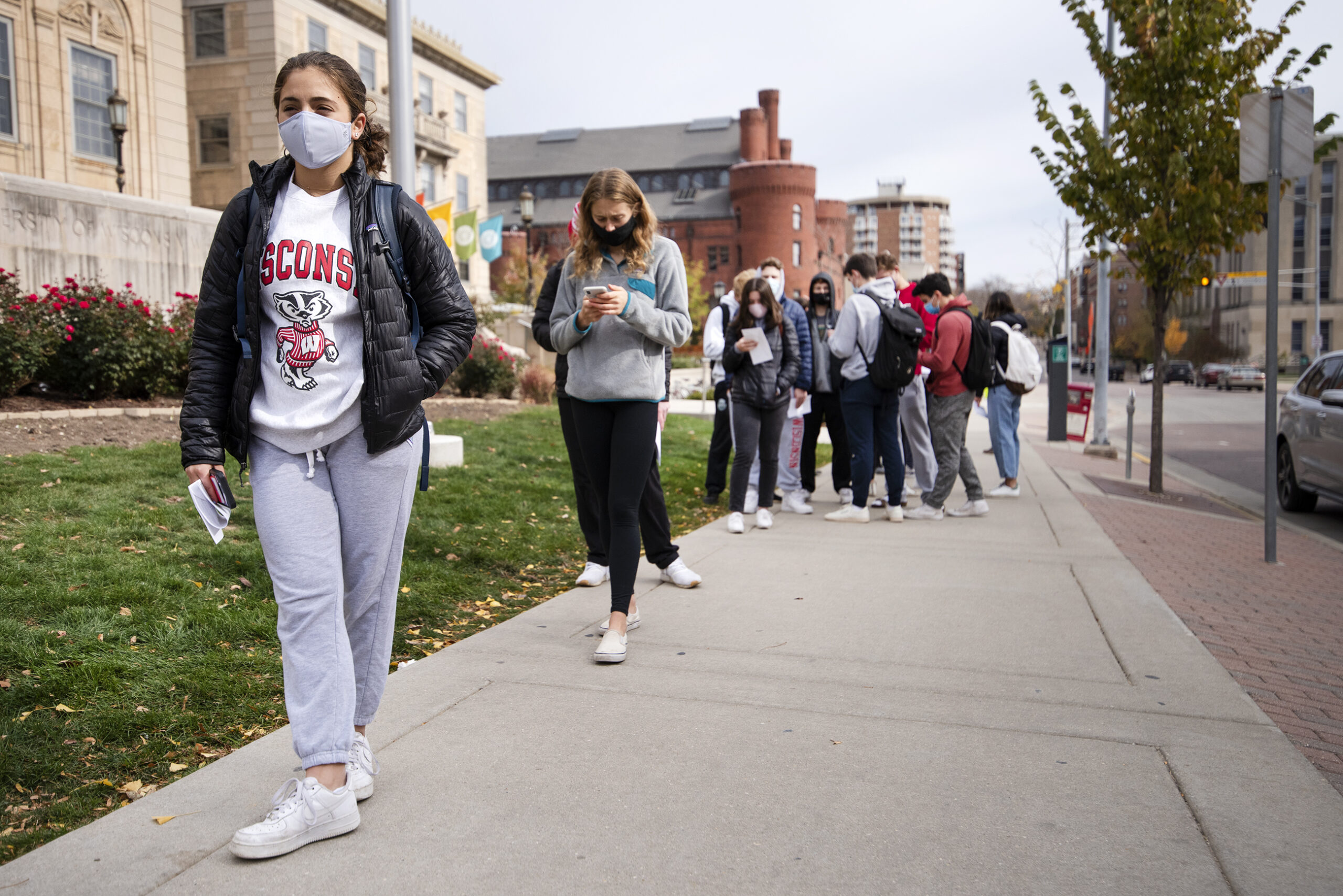 A student in a face mask and Bucky Badger t-shirt holds a ballot while waiting in line on a sidewalk