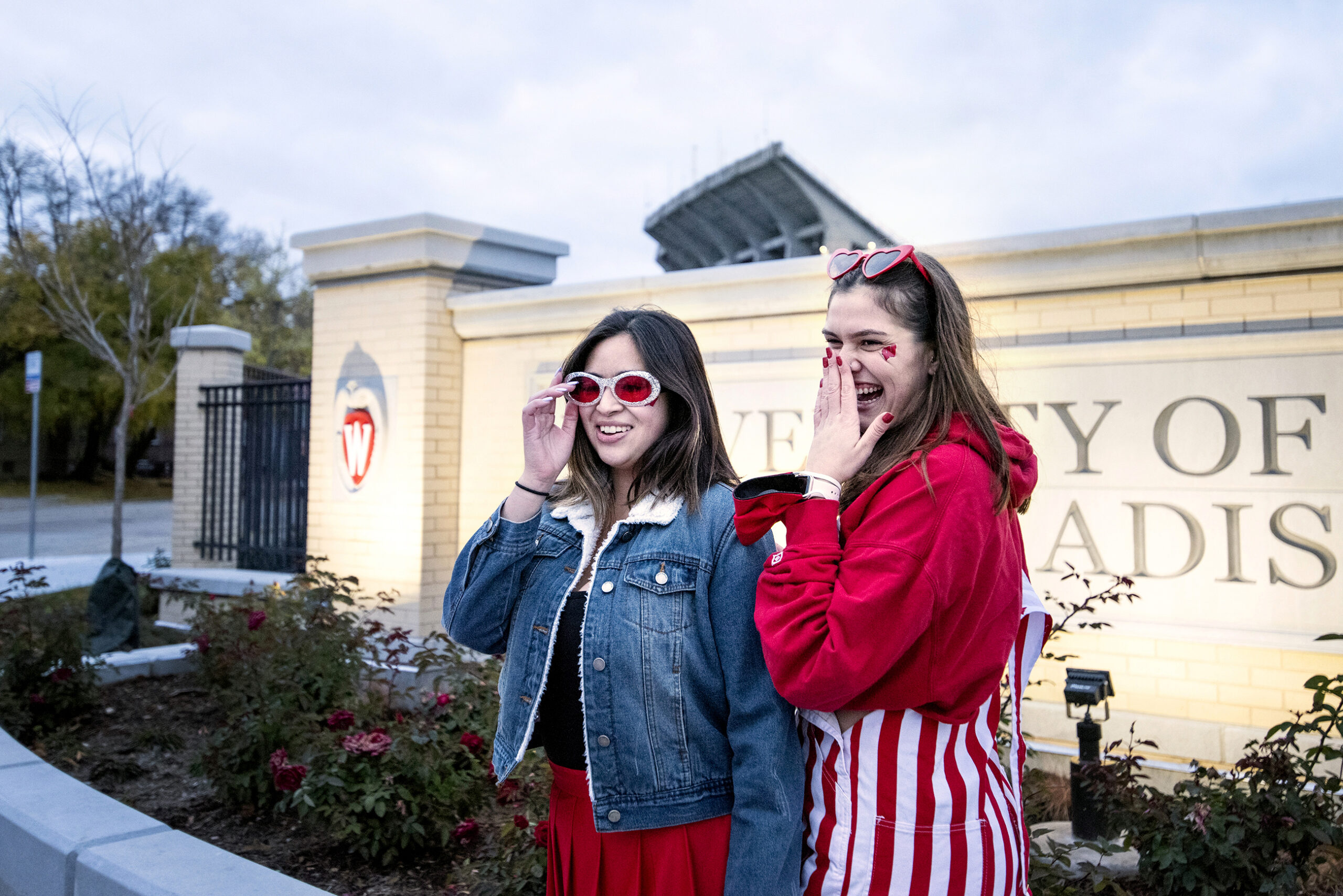 girls stand in front of a sign at Camp Randall in badgers gear and sunglasses