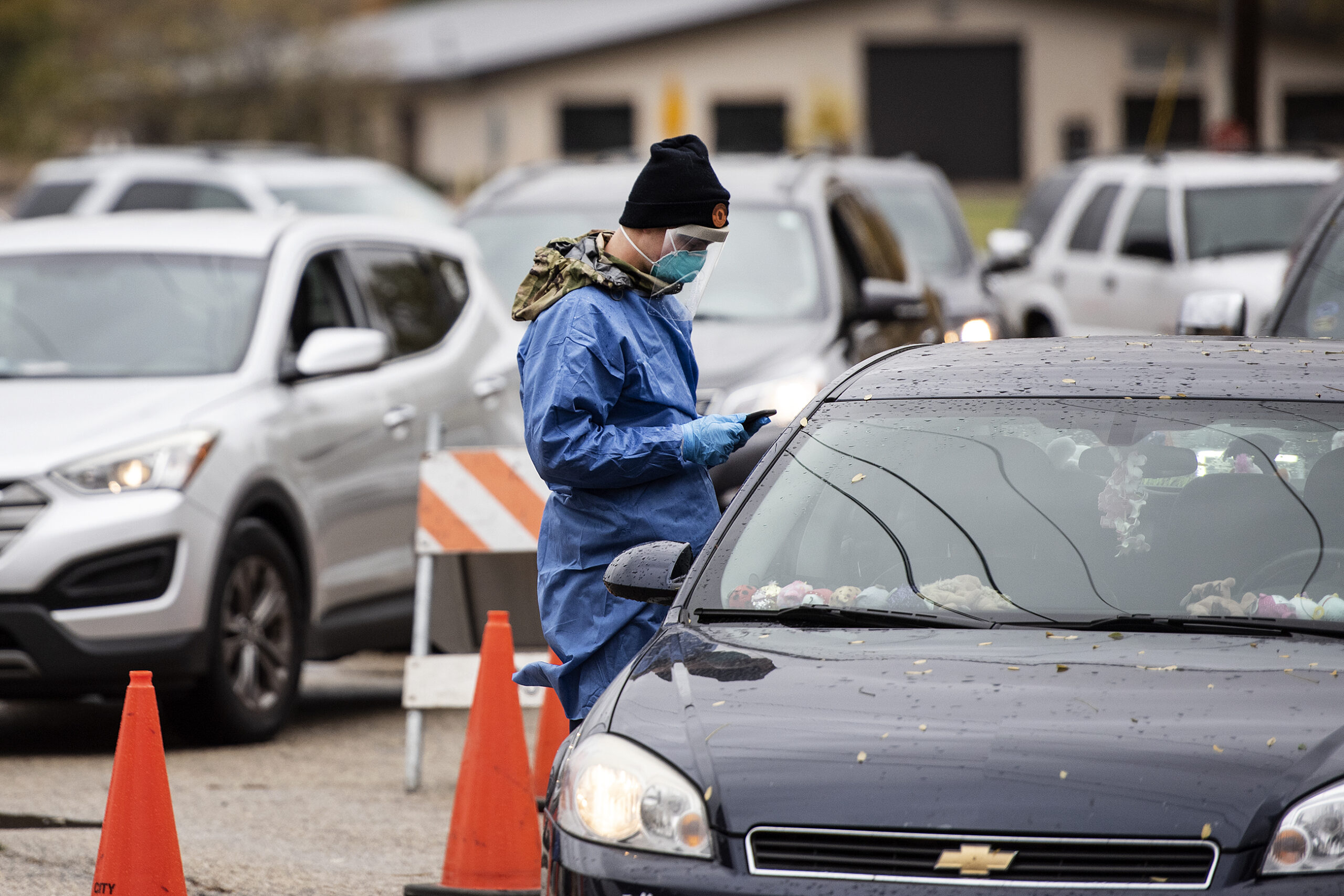 a man in a blue gown and face covering uses a phone as he approaches a vehicle in line for the test