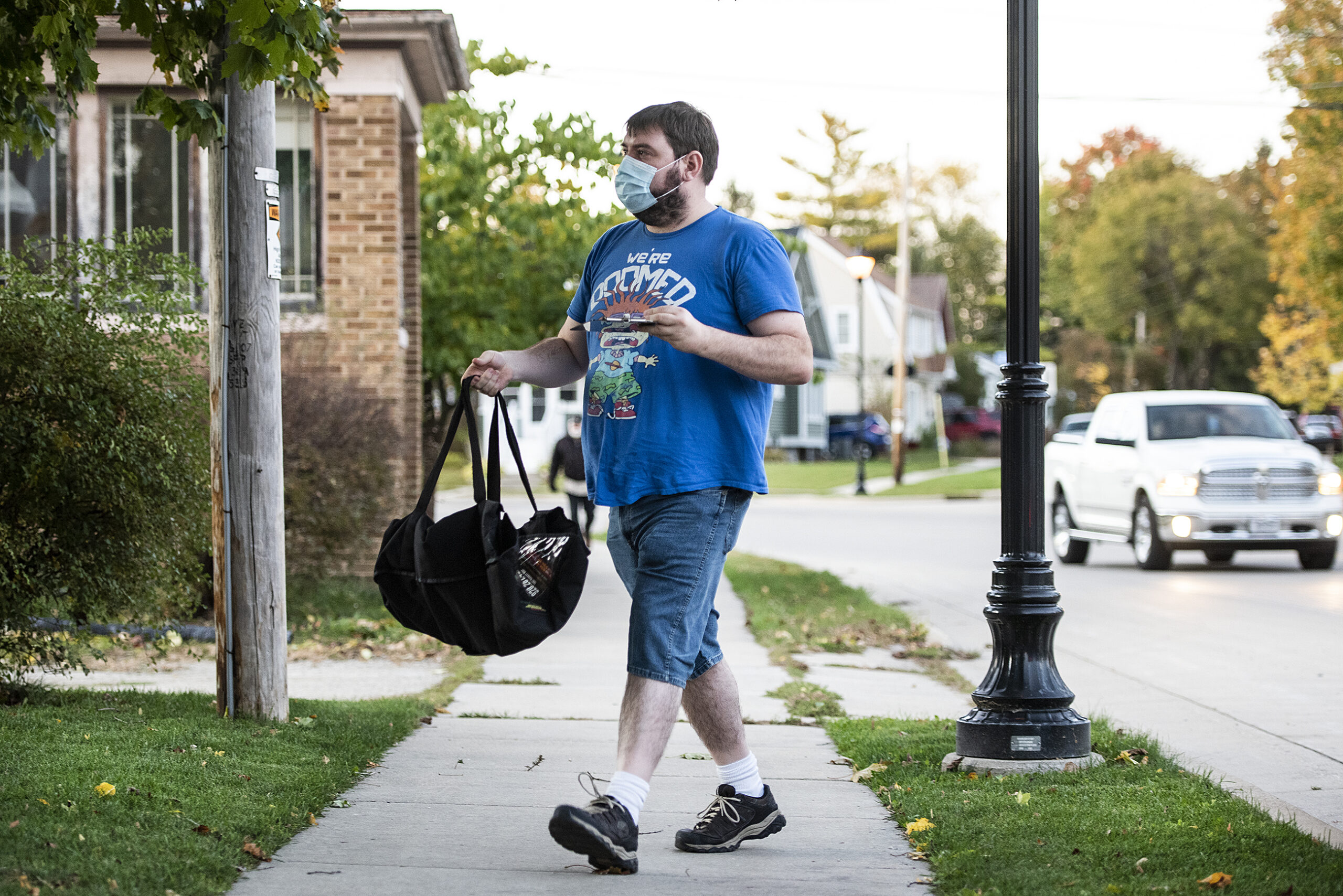 a man in a face mask walks across a sidewalk as he carries a black bag containing a pizza