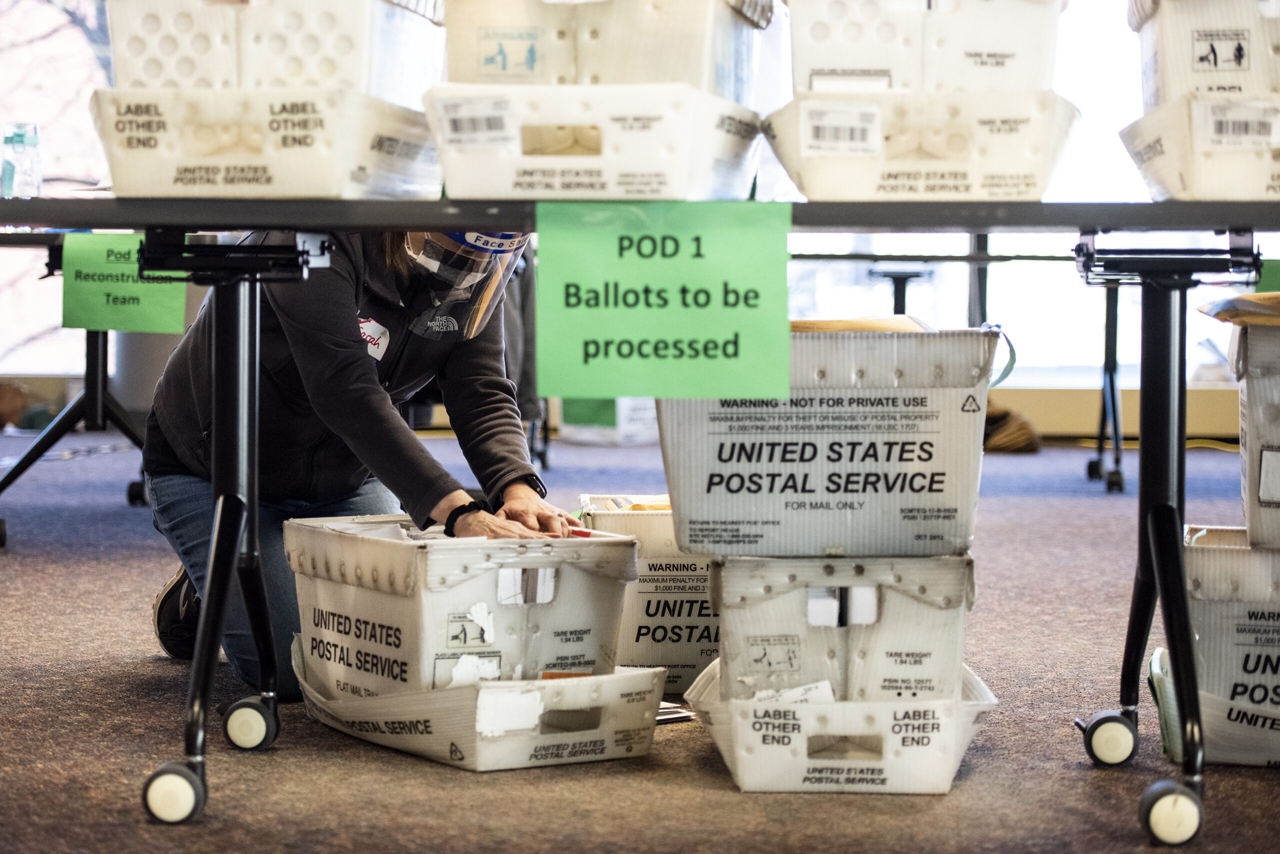 crates of ballots are on the floor and tables as a worker in a face shield flips through them with her hands