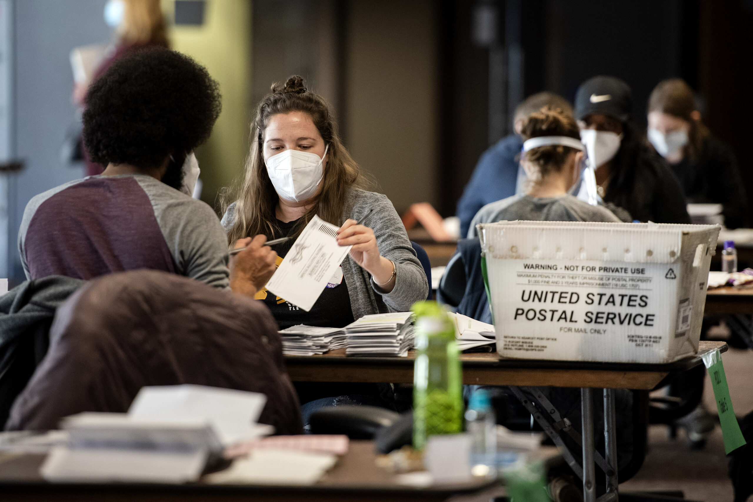 a worker in a white face mask holds a ballot while sitting at a table
