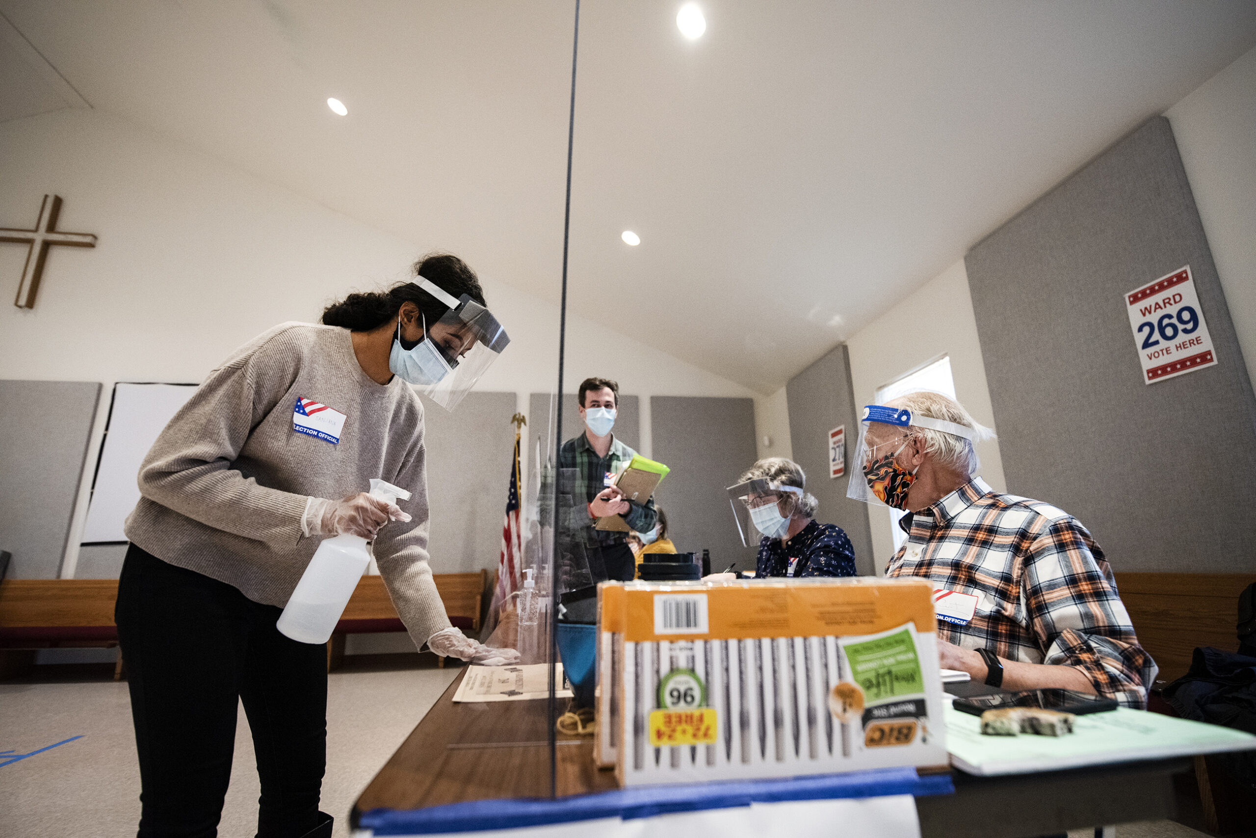 A woman in a mask and face shield holds a spray bottle and wipes a table with a cloth as poll workers sit across from her on the other side of plexiglass