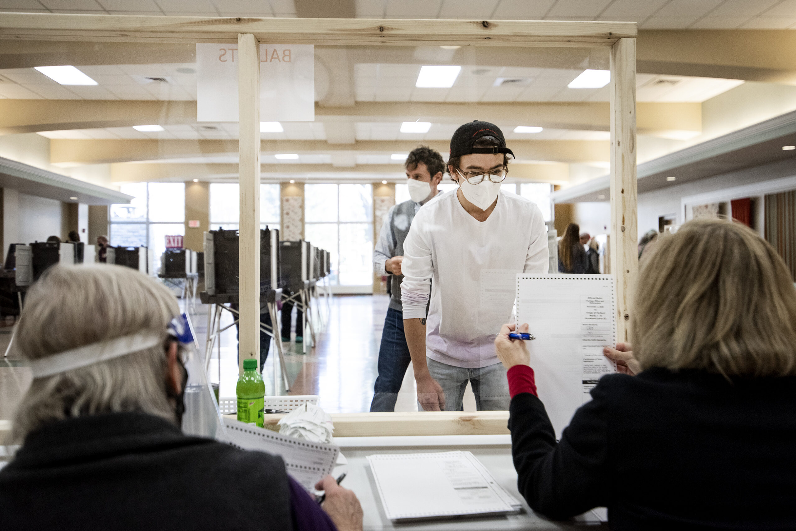 A voter in a white face mask looks through plexiglass as a poll worker holds up a ballot