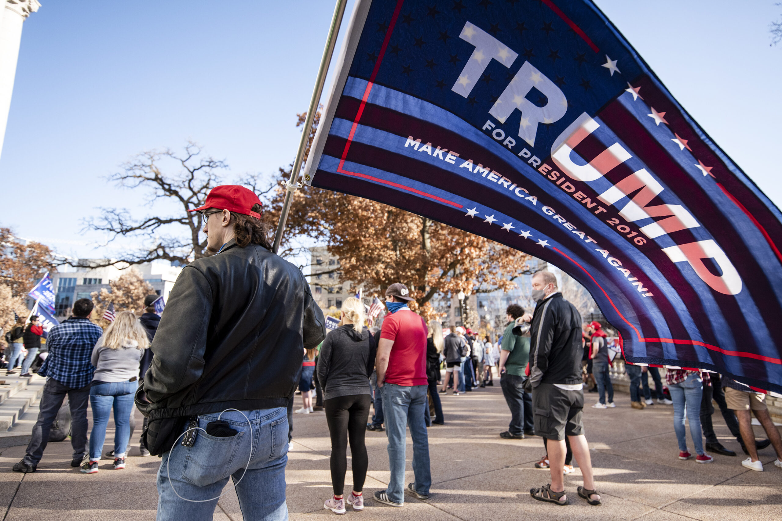 A man holds a Trump flag that waves in the wind. Supporters gather around in front of the capitol.