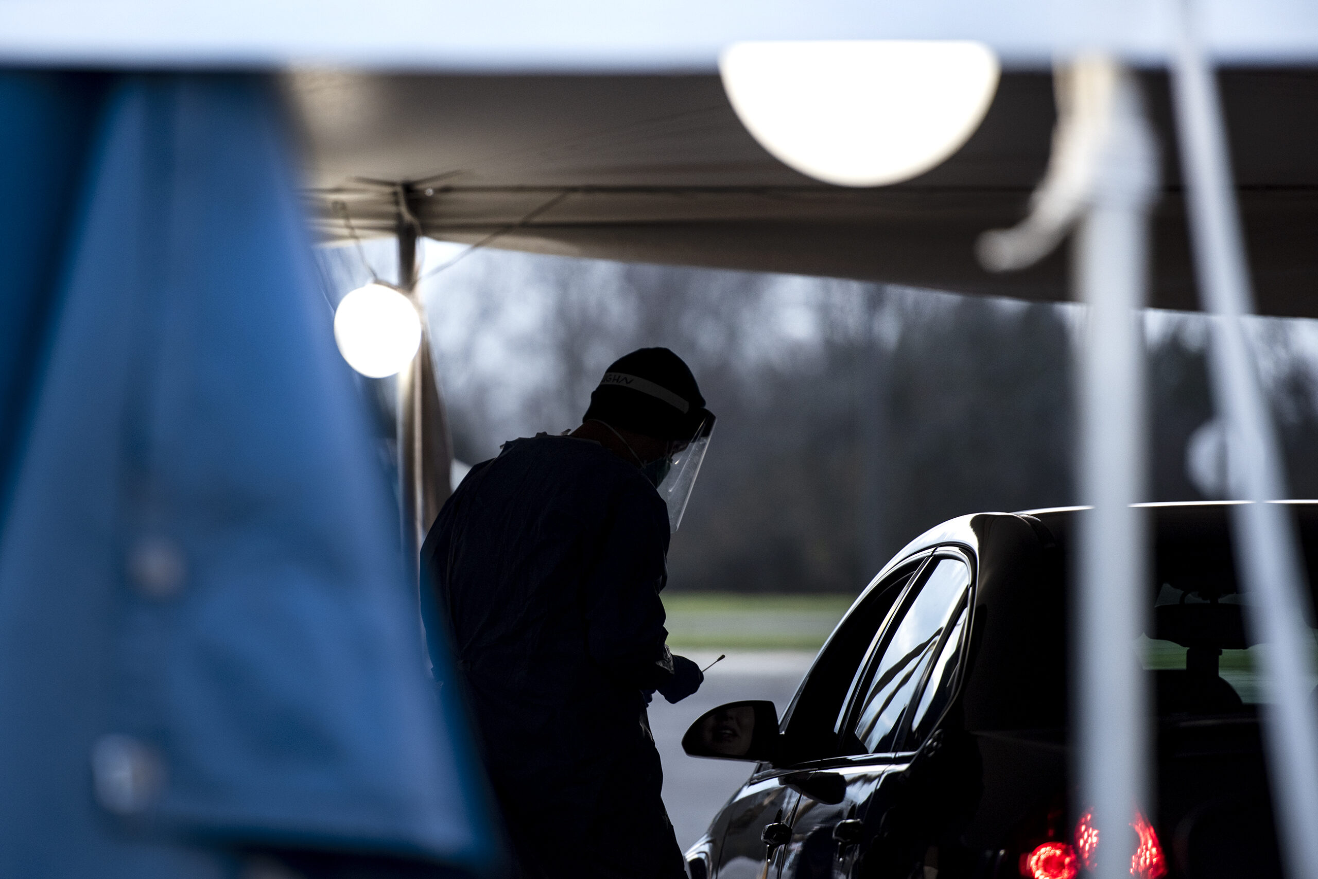 A man in a face mask holds a swab as he approaches the driver of a vehicle. The man is in silhouette and under a tent as the sun begins to set.