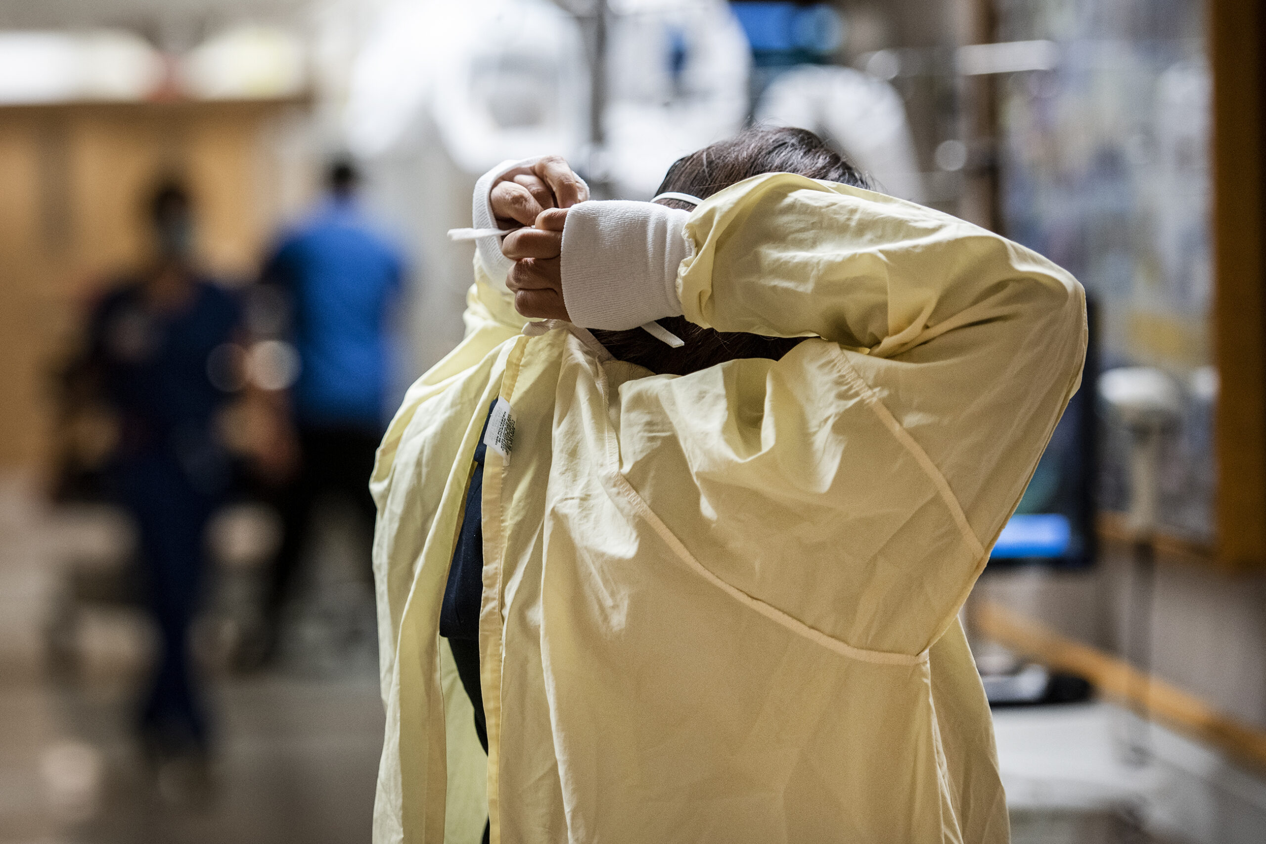 A woman can be seen reaching behind her head to tie up a yellow medical gown