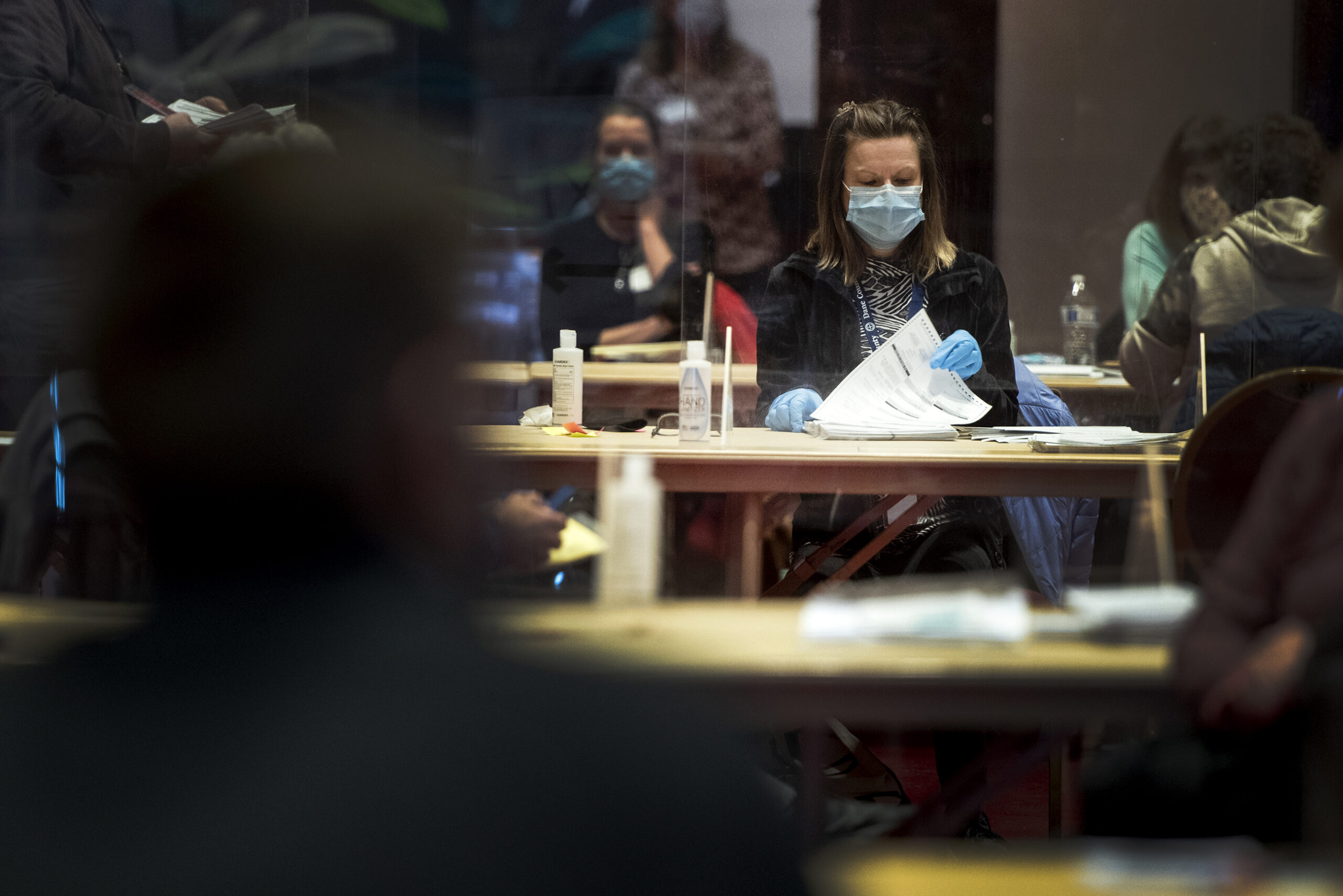 Lines of tables can be seen as a worker in gloves and a face mask flips through ballots