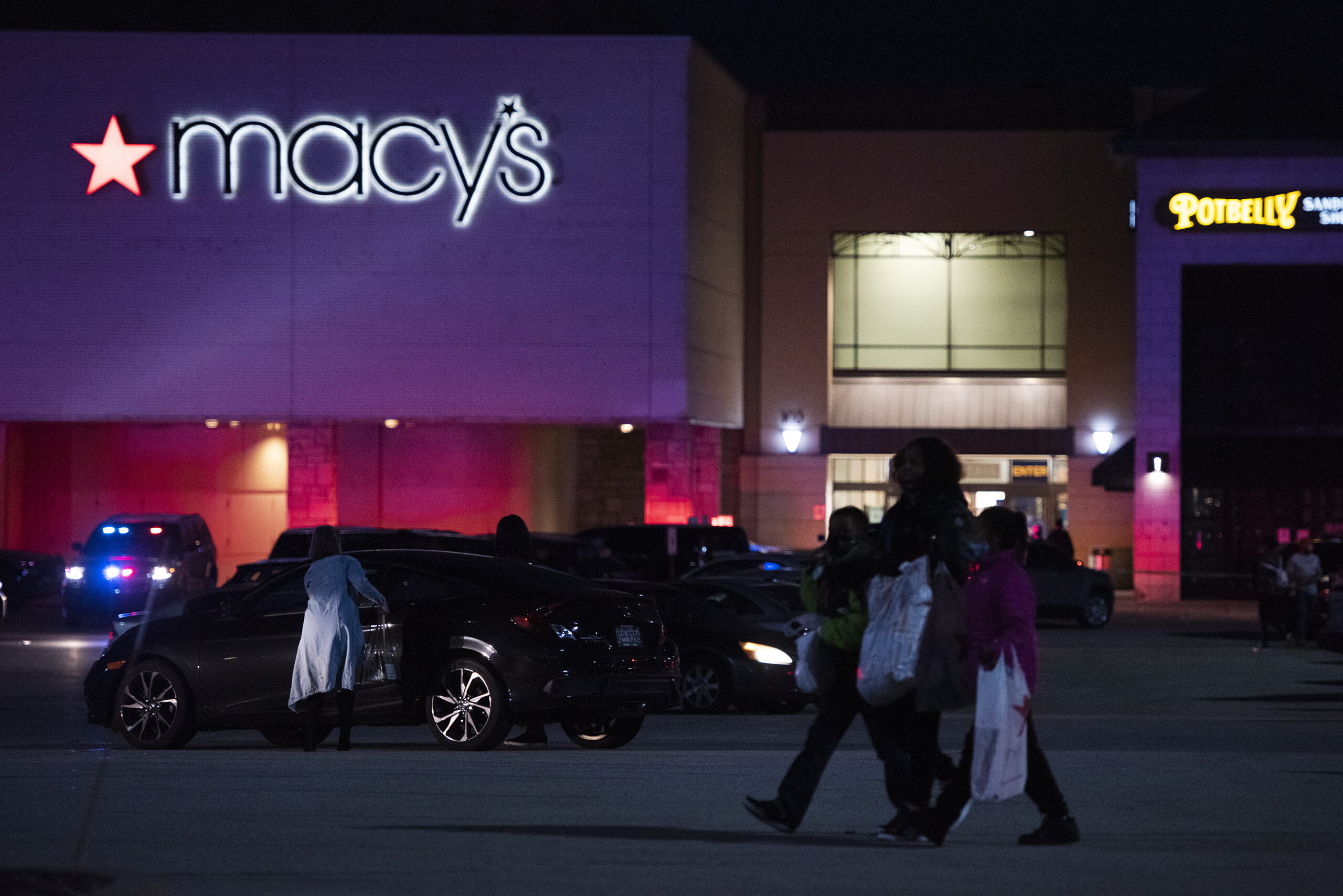 Three people with white shopping bags pass through a dark parking lot in front of a Macy's