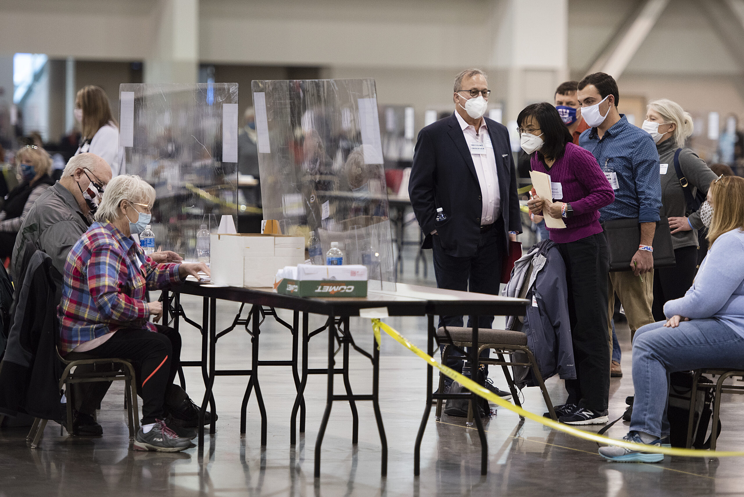 Several election observers lean forward to peer through plexiglass at two people counting ballots