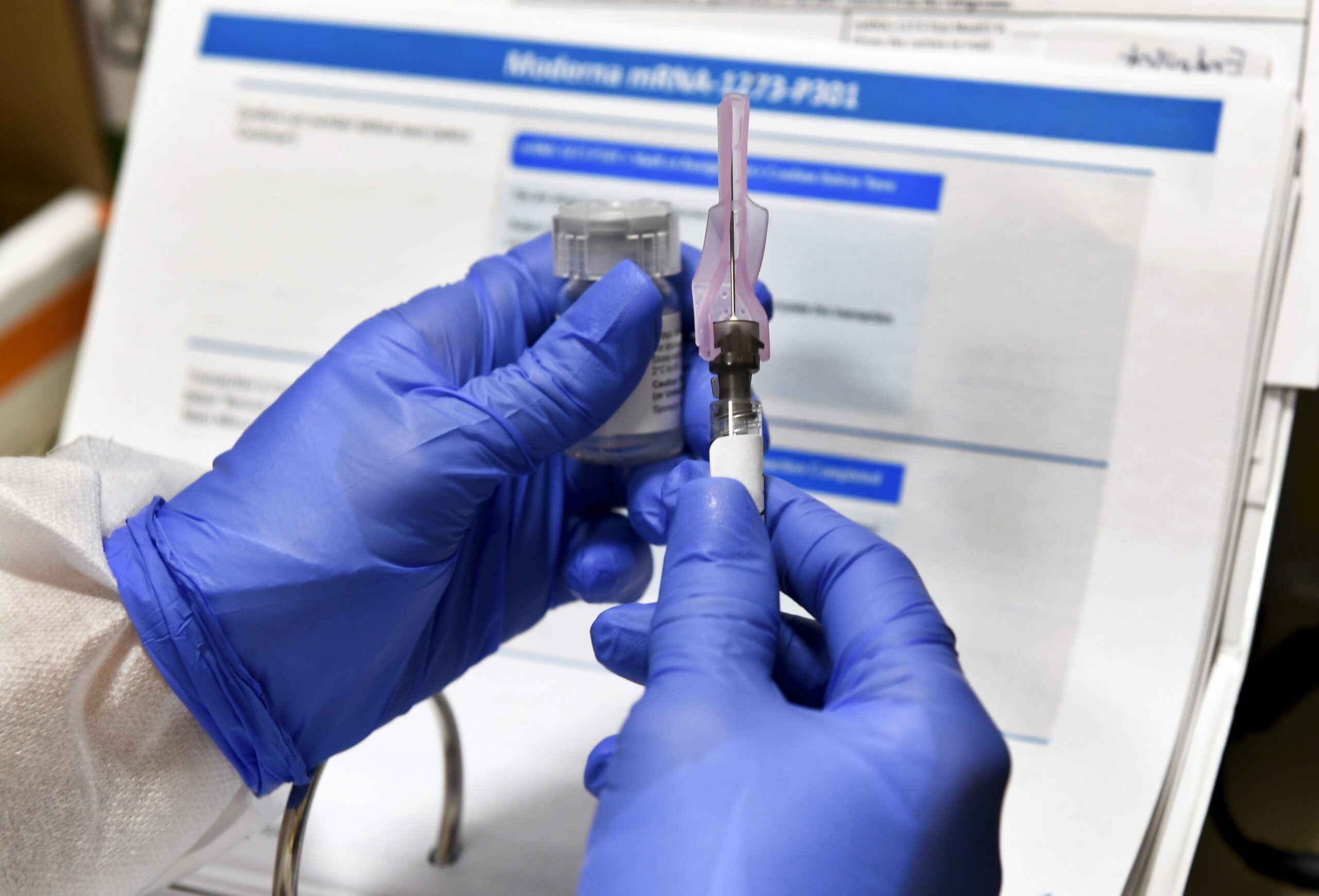 Two hands with blue gloves hold a syringe of fluid being tested as the COVID-19 vaccine