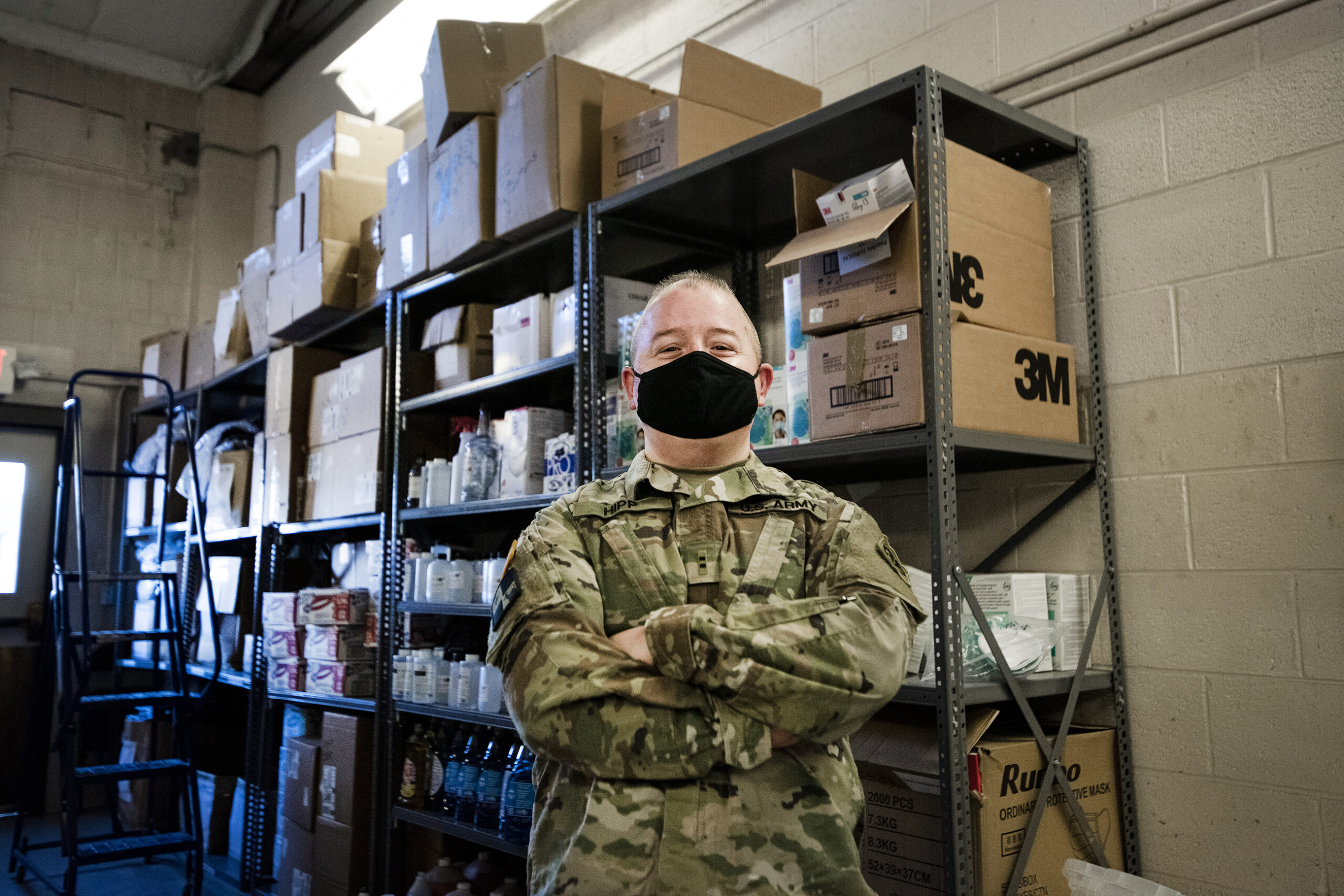 a man in a military uniform and a black face mask stands near shelves of PPE with his arms crossed