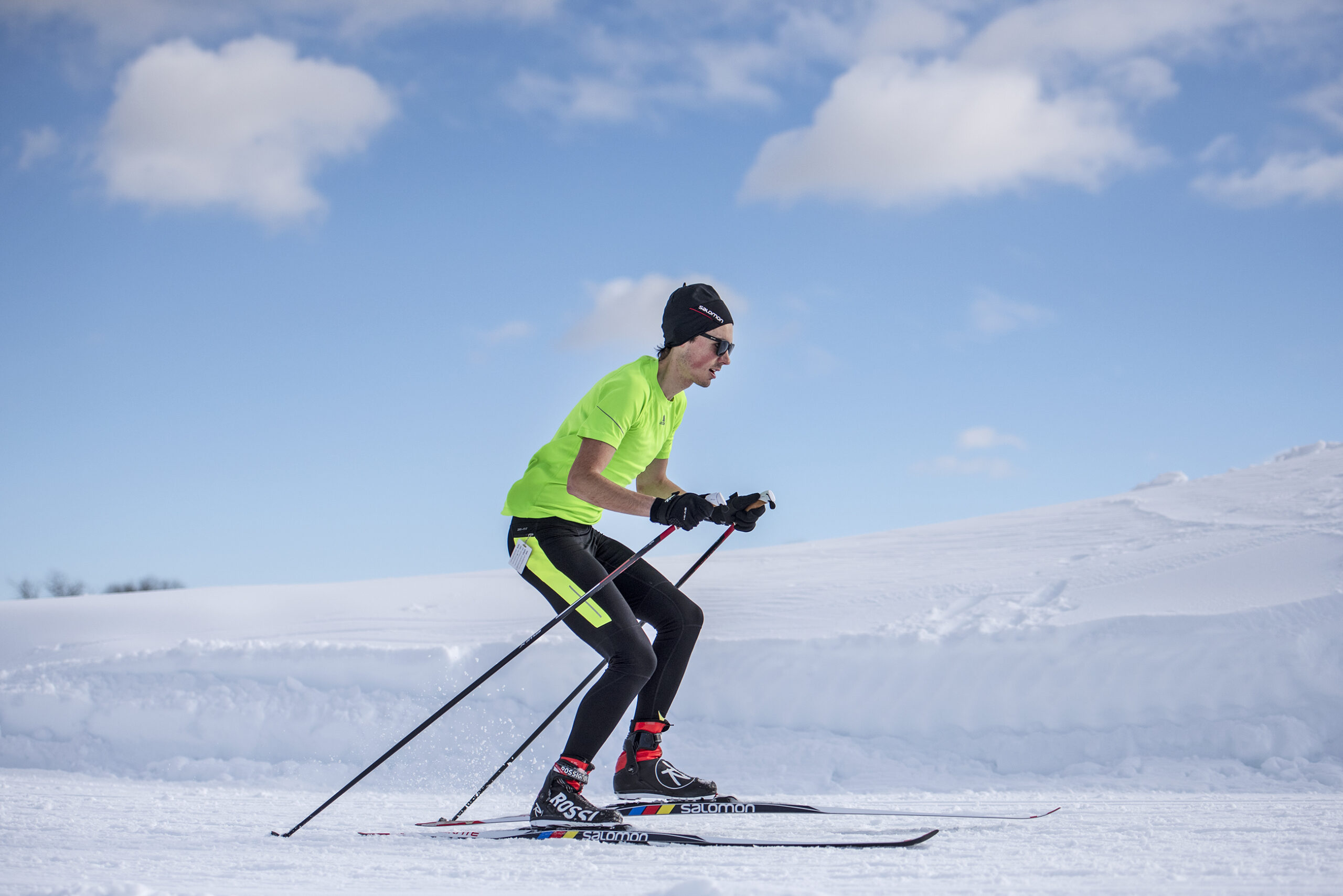 A man in a neon yellow short-sleeve shirt skis on white snow under a blue sky
