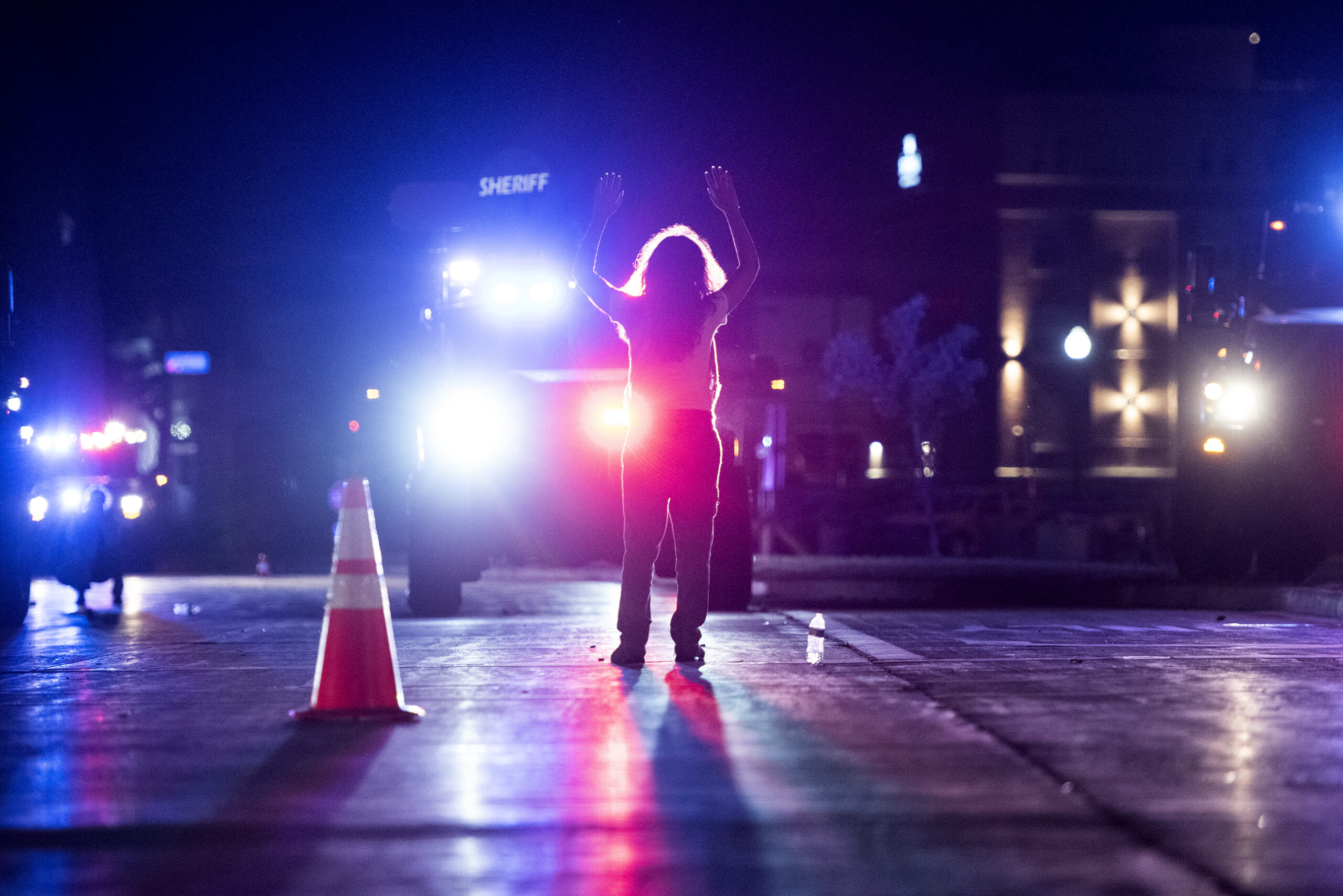 blue and red lights fill the night as a lone protester raises their arms in front of large law enforcement vehicles