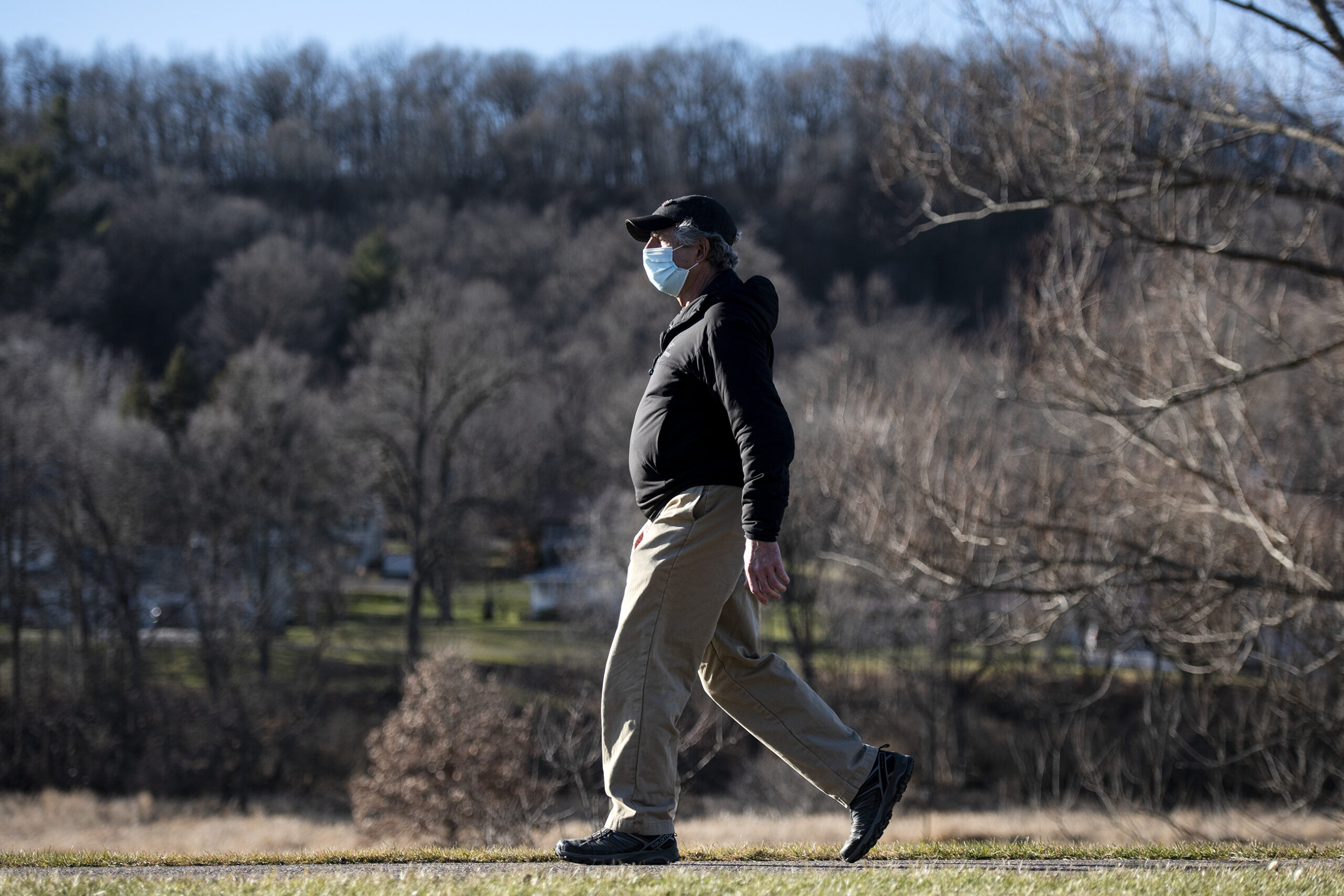 Scenery can be seen behind a man walking outside in the sunshine. He wears a face mask.