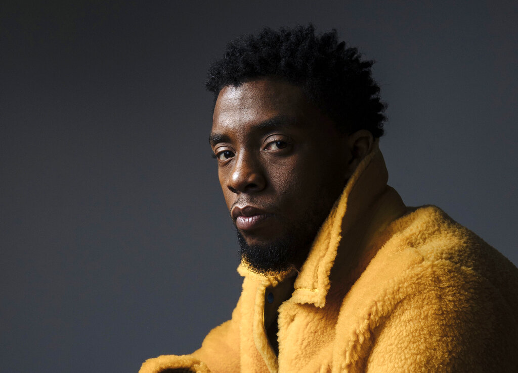 A close-up photo of actor Chadwick Boseman is shown wearing a plush-like golden yellow jacket against a black background looking directly into the camera.
