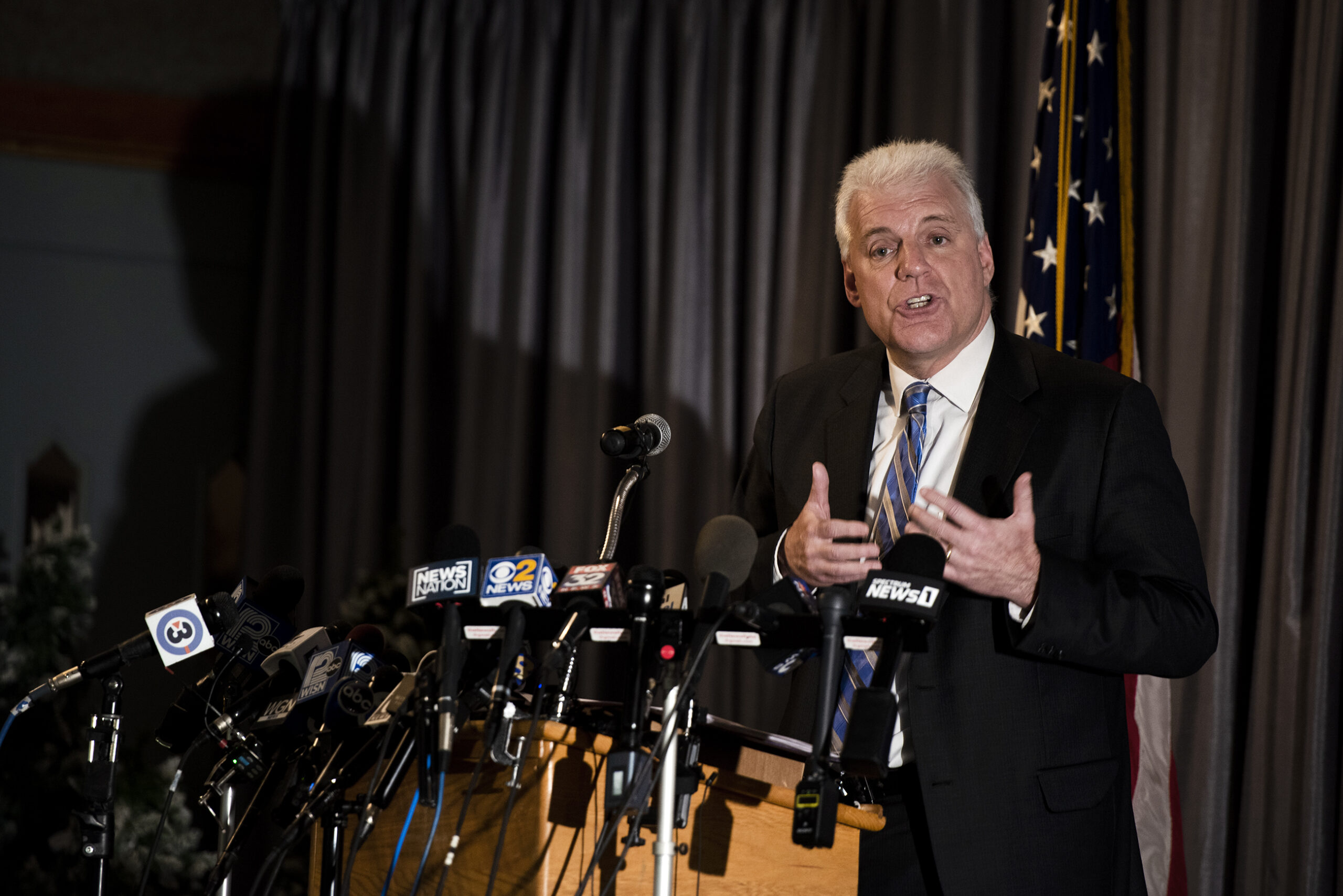 A man in a suit gestures as he speaks to reporters from a stage