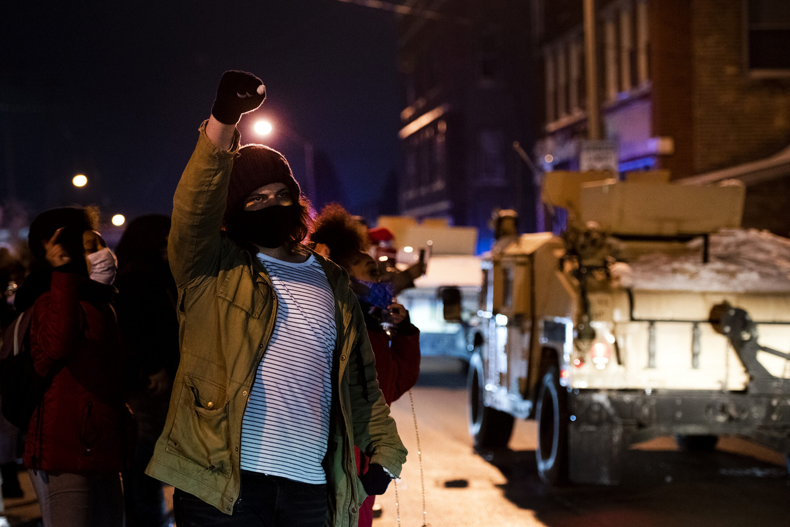 military style vehicles drive by as a protester raises a fist