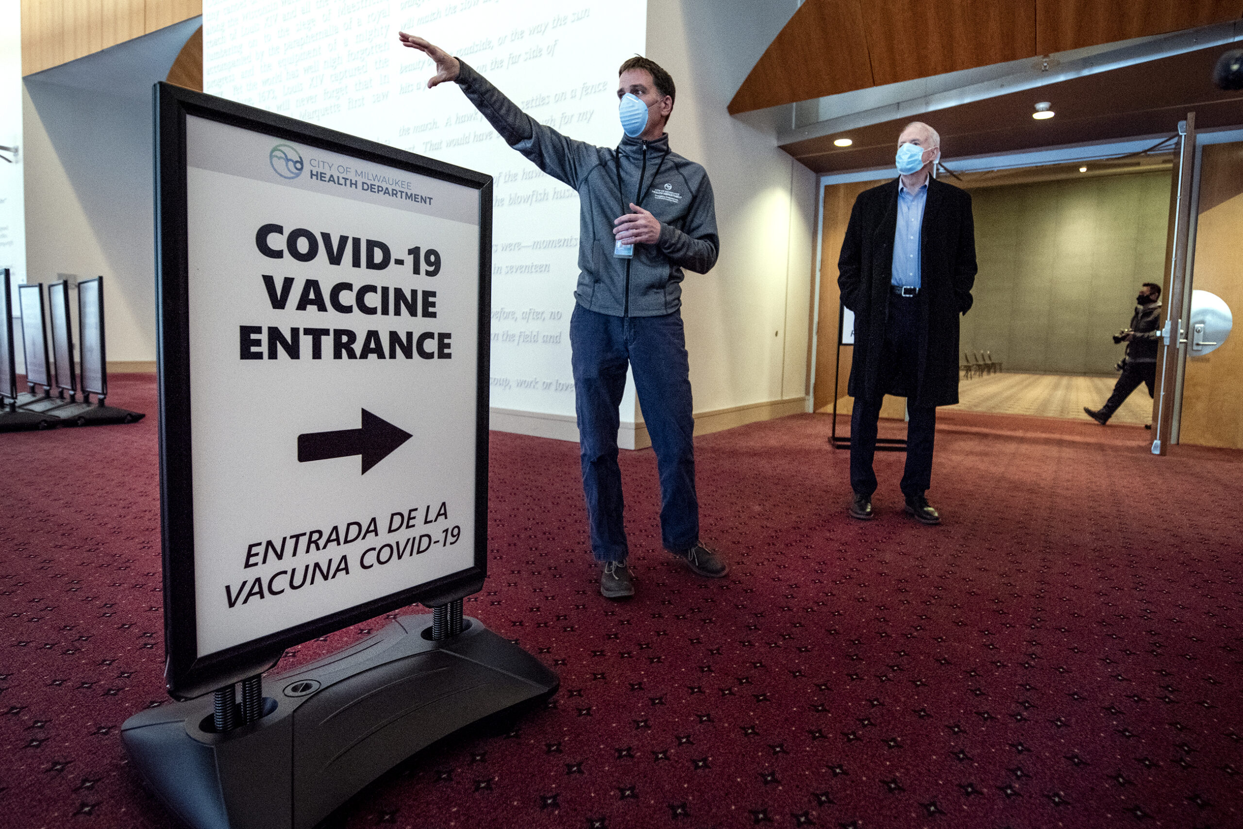 Two men in face masks stand by a sign that says "COVID-19 Vaccine Entrance"