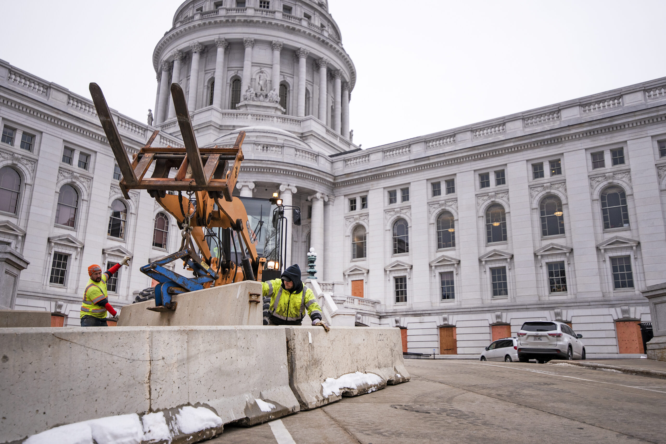 Two workers in yellow jackets stand in front of a construction vehicle as they place concrete barriers in front of the state capitol