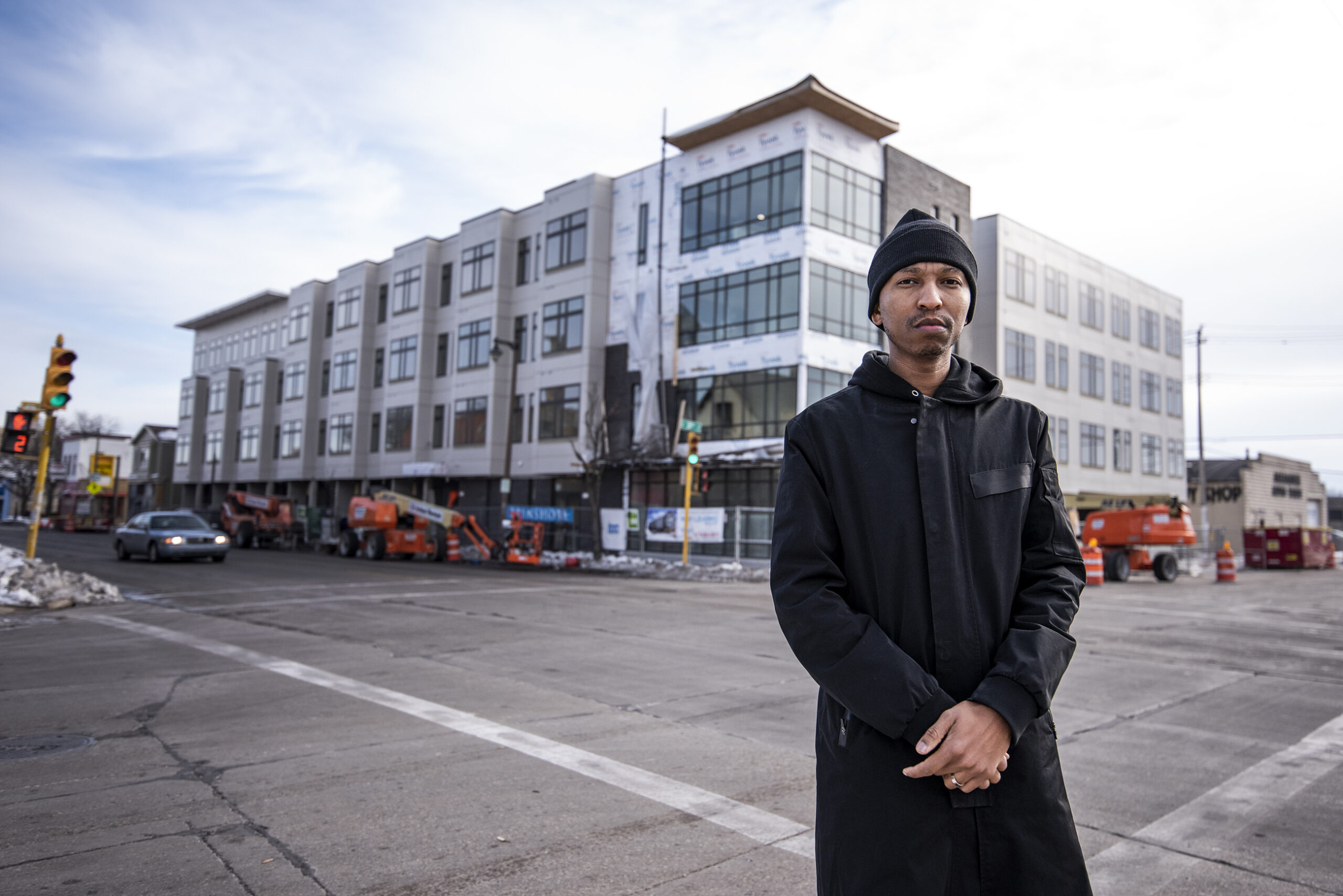 A man in a black coat stands in front of an apartment that is under construction, but mostly complete.