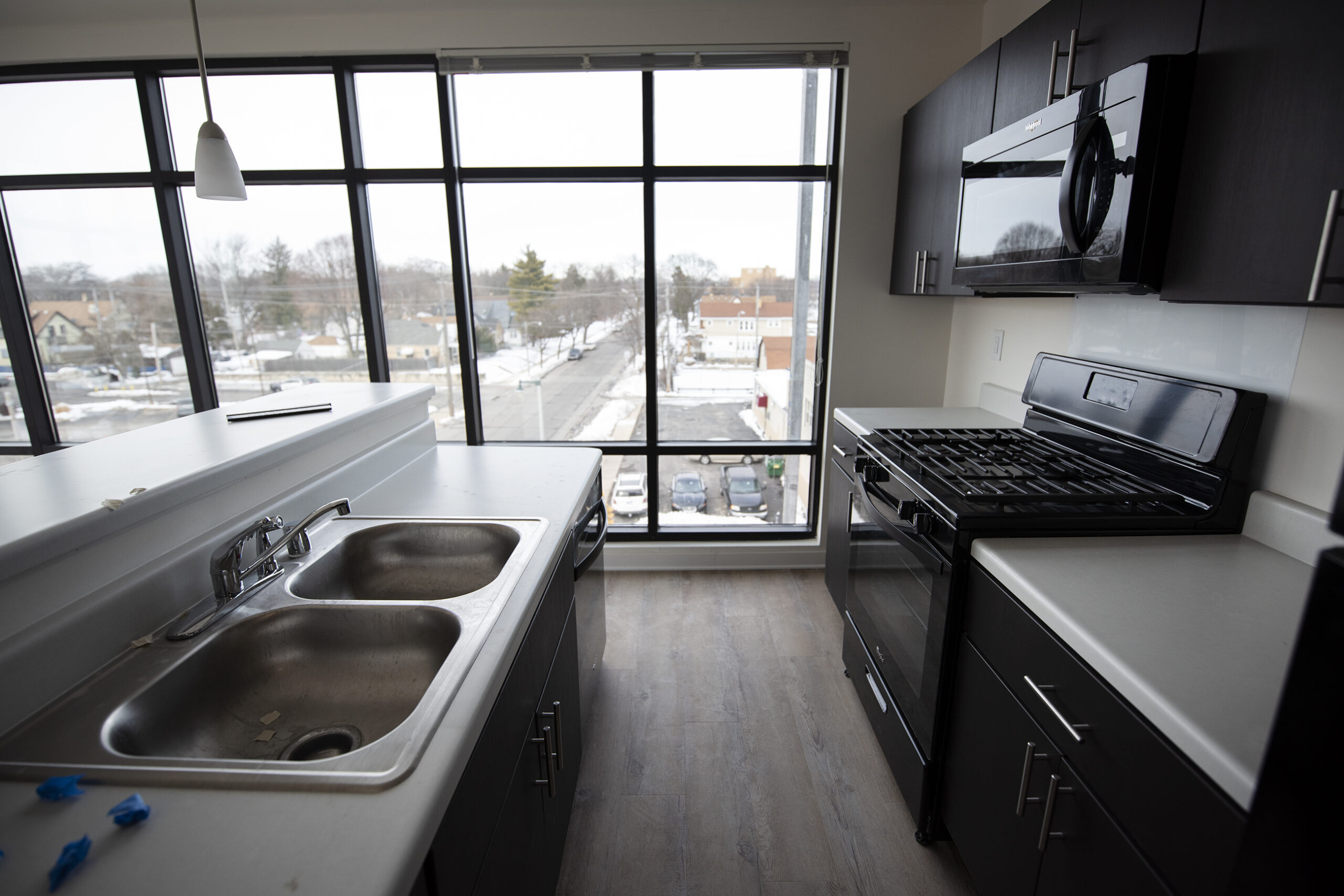White countertops and black appliances create a small kitchen area that is surrounded by large windows taking up nearly the entire wall