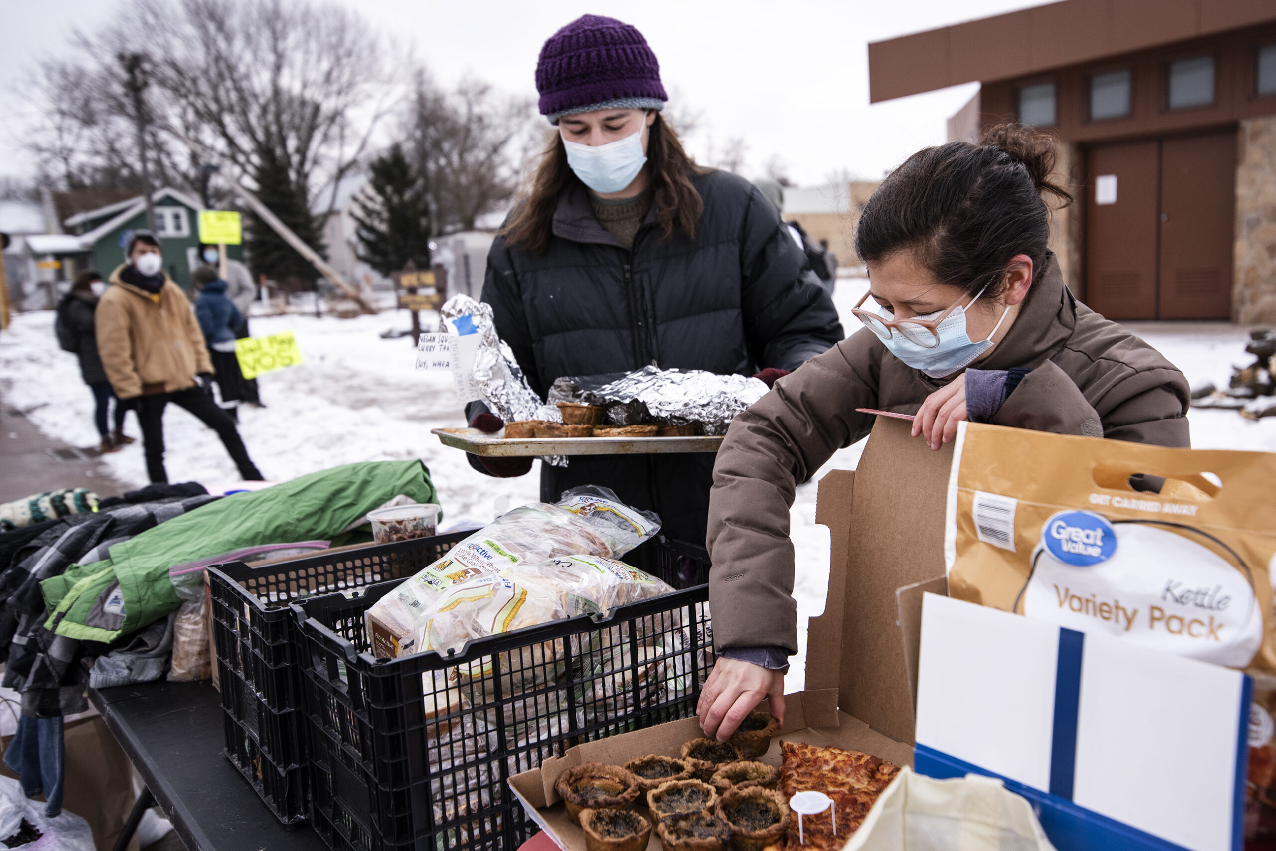two people in winter gear and face masks place small tarts into a box next to other donated items