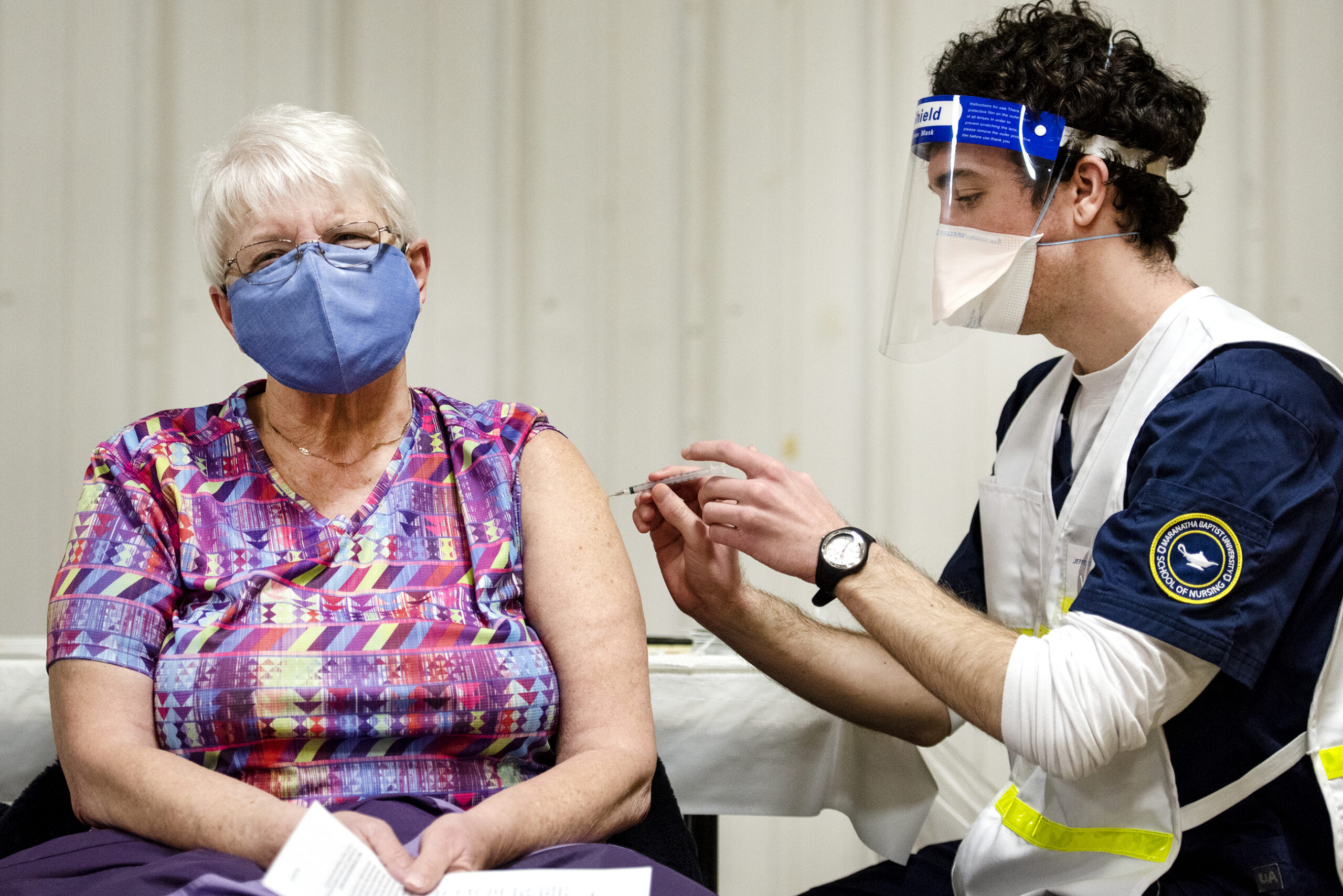 a woman in a purple mask has a rolled up sleeve as a student nurse gives her the covid-19 vaccine