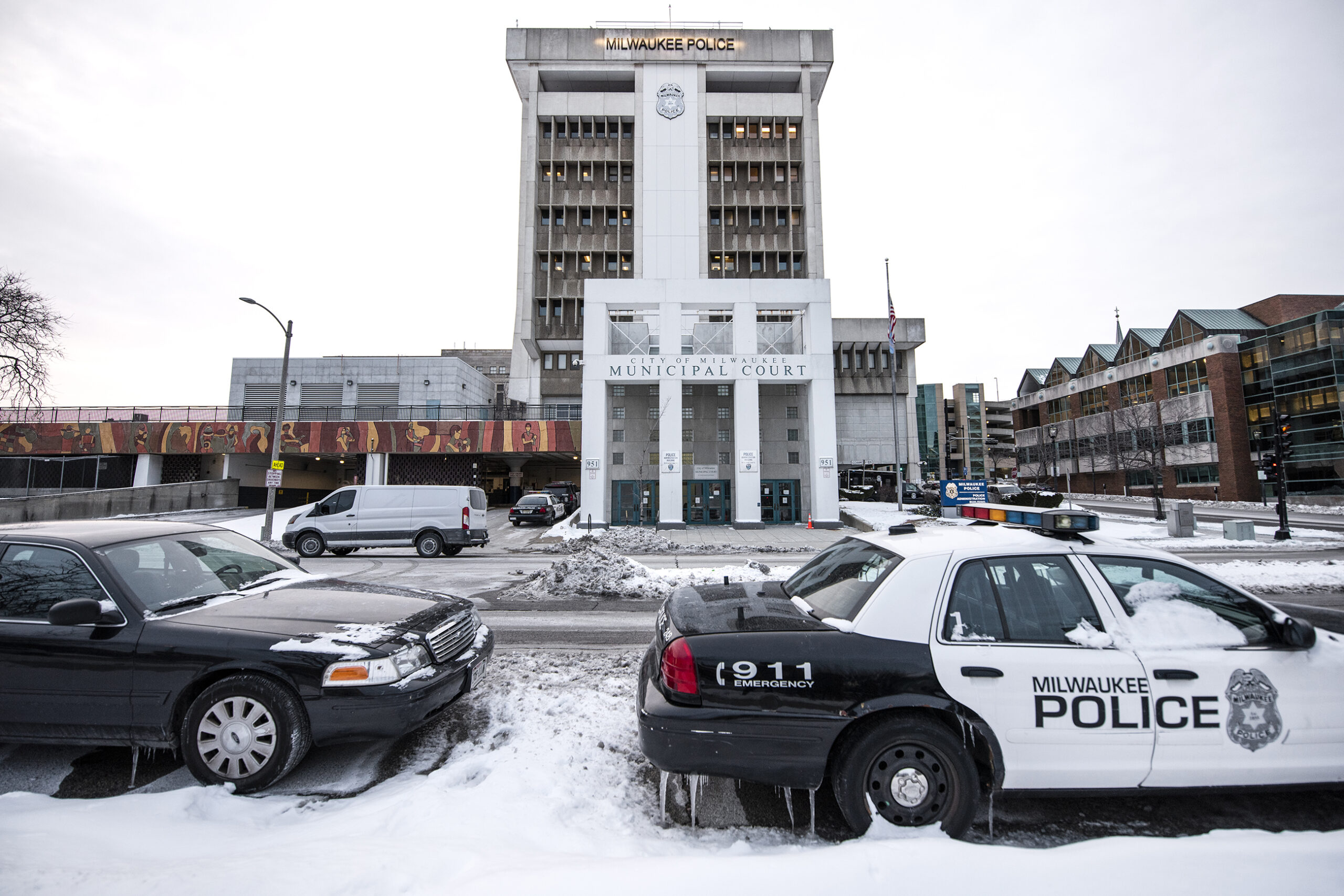 Two Milwaukee police vehicles are parked on a snowy street. The Milwaukee police department is in the background.