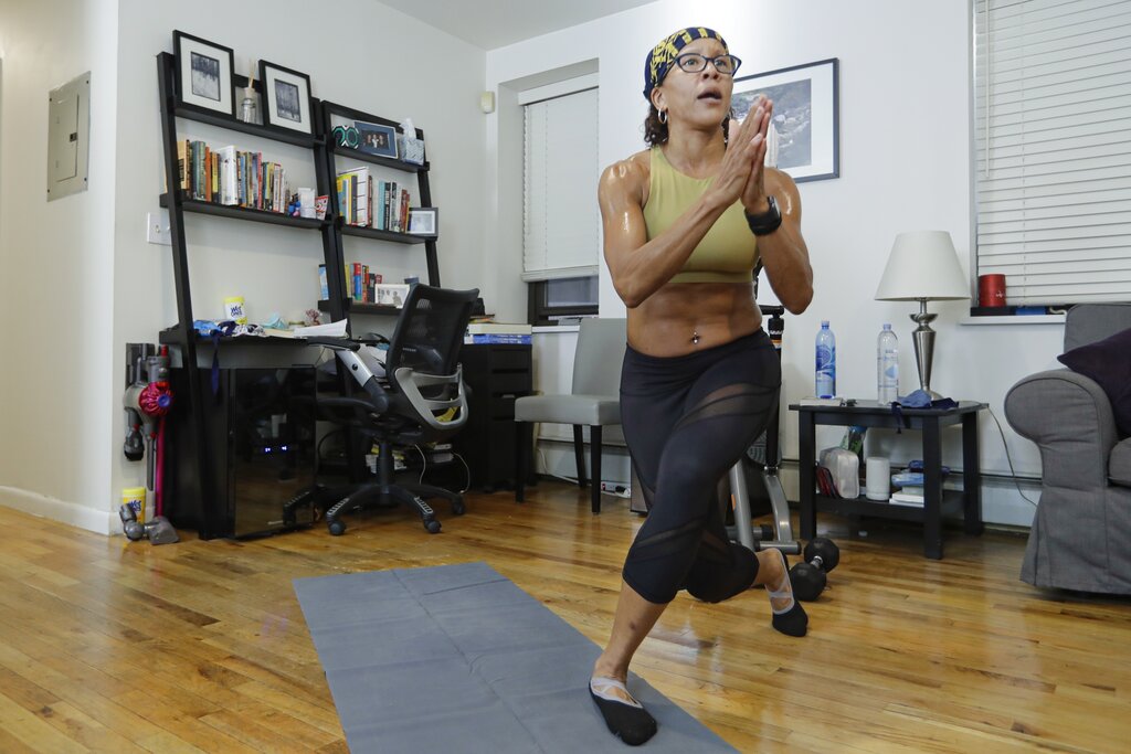 A woman does a lunging pose on her yoga mat in her home with a computer desk setup in background.