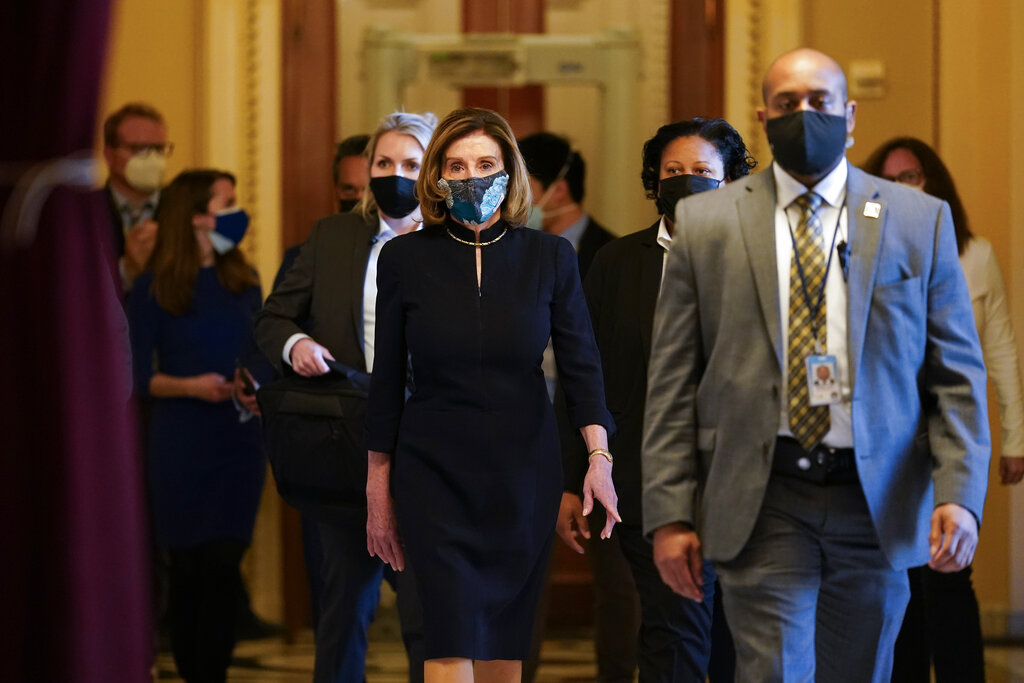 Nancy Pelosi walks down a hallway wearing a mask with others gathered in the hallway around her.