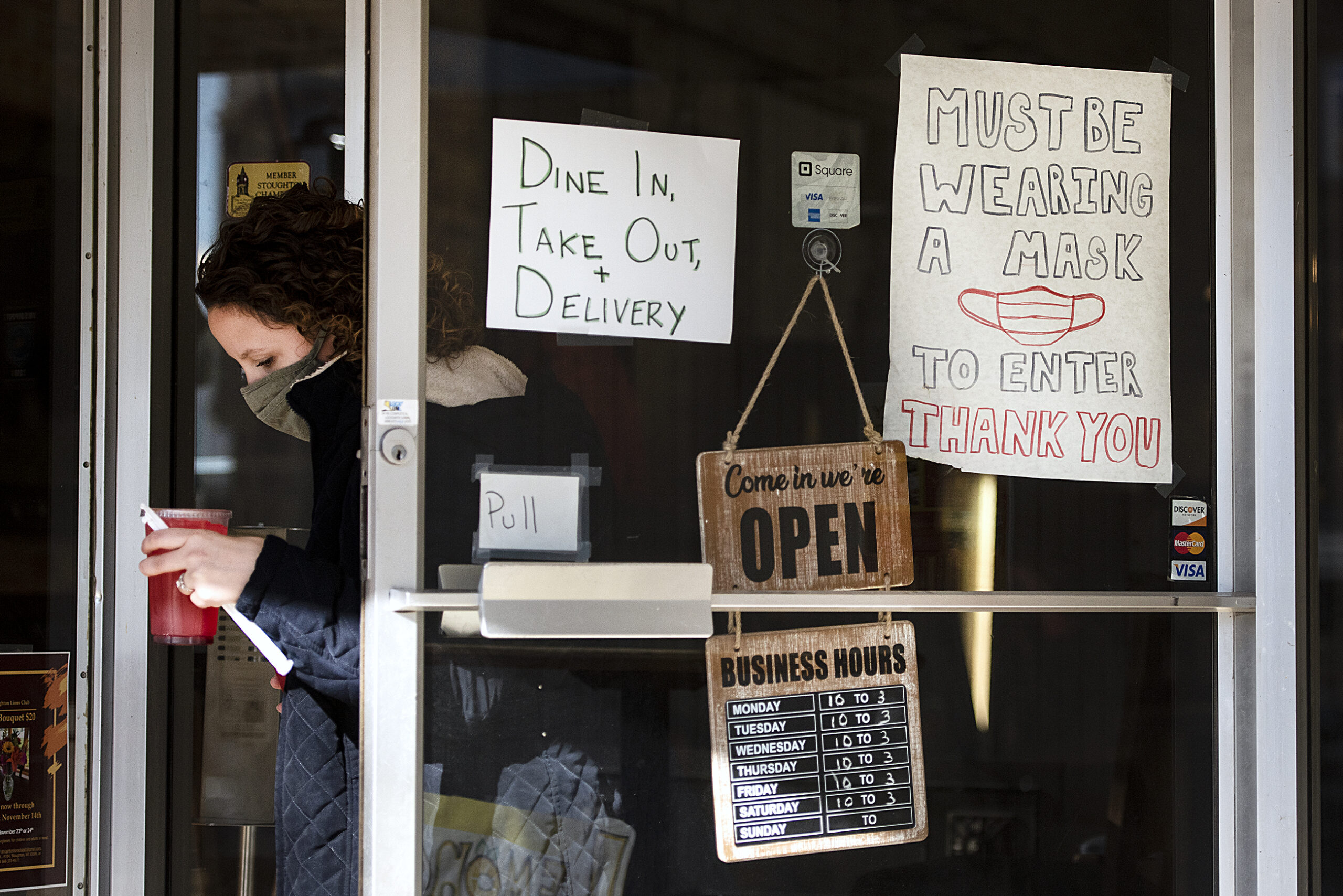 A woman in a face mask holds a drink as she pushes open a door that has signs indicating mask requirements for entry