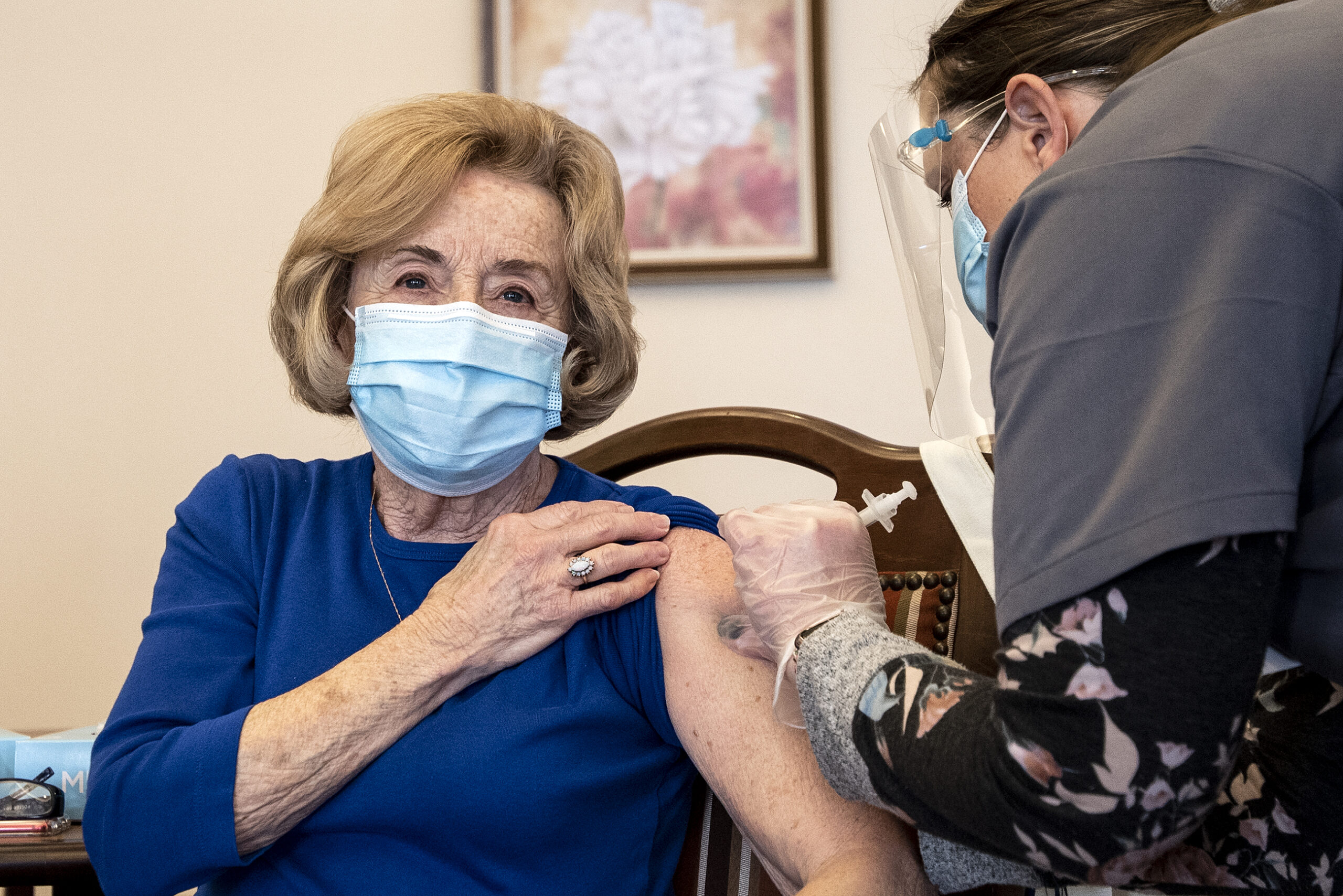 A woman in a blue shirt and blue face mask holds up a sleeve as she receives a shot