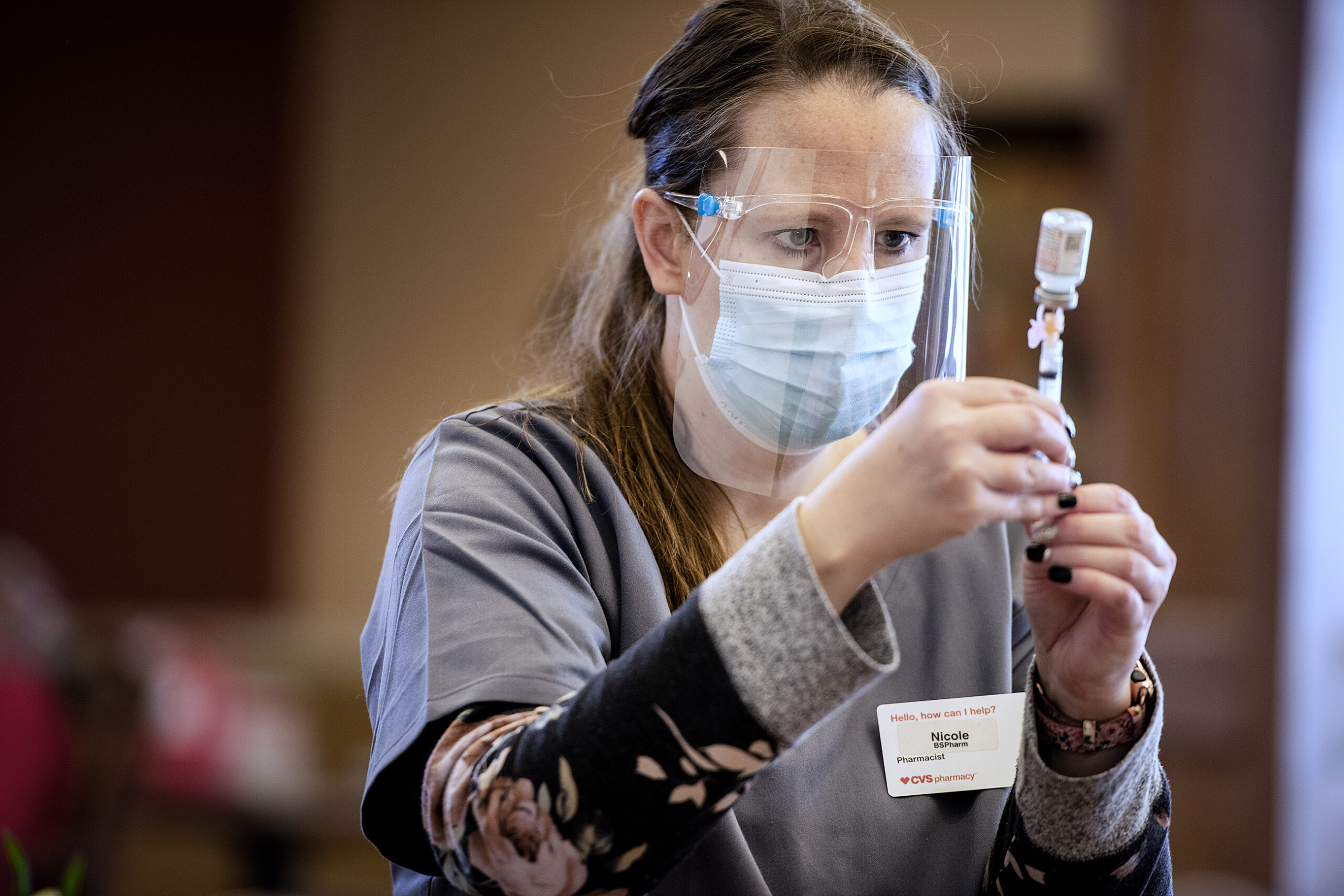 A pharmacist in gray scrubs and a face shield looks closely at a vial as she draws up a vaccine