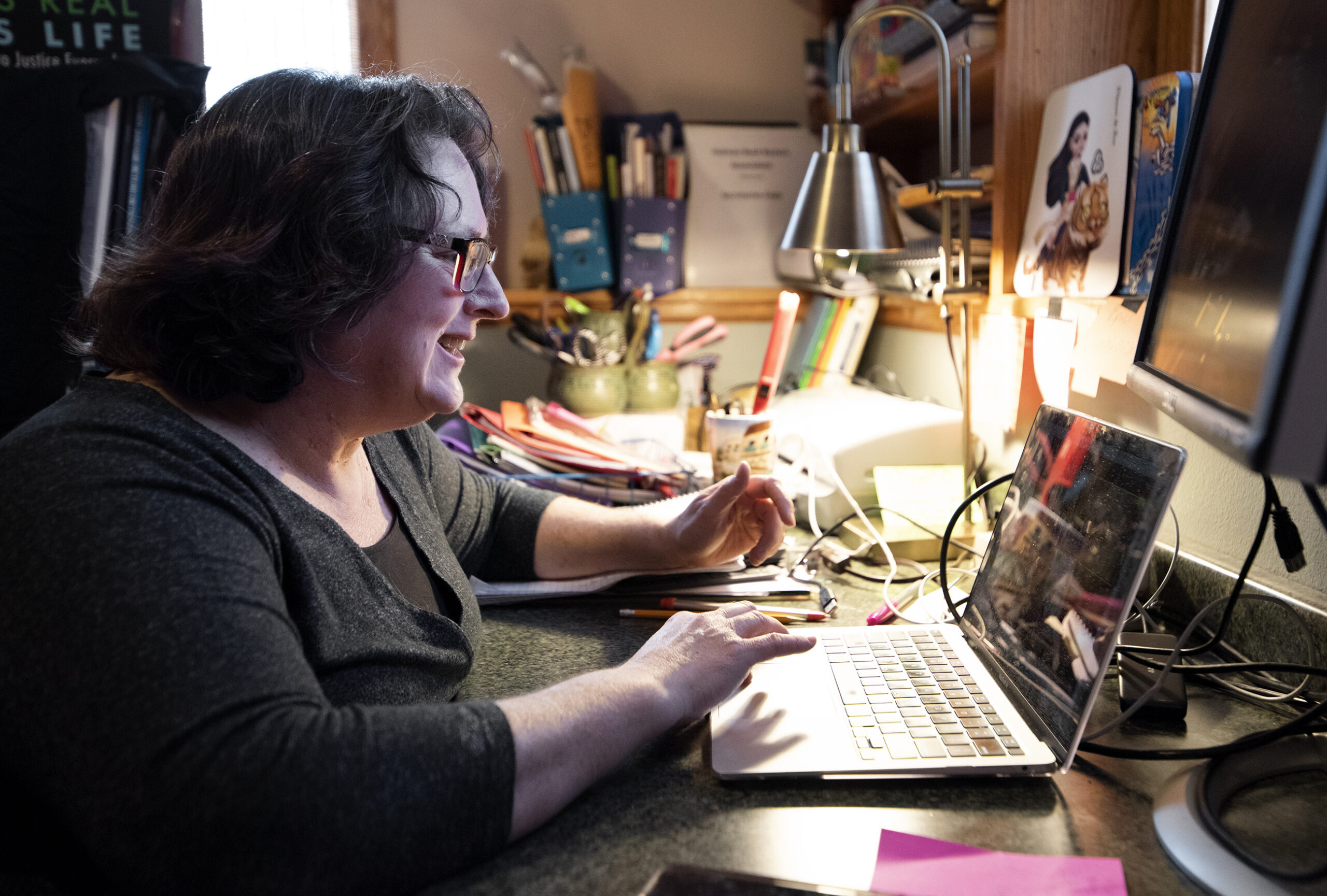 a woman sits in front of a laptop as she speaks to students. A desk lamp and various pictures surround her at her desk.
