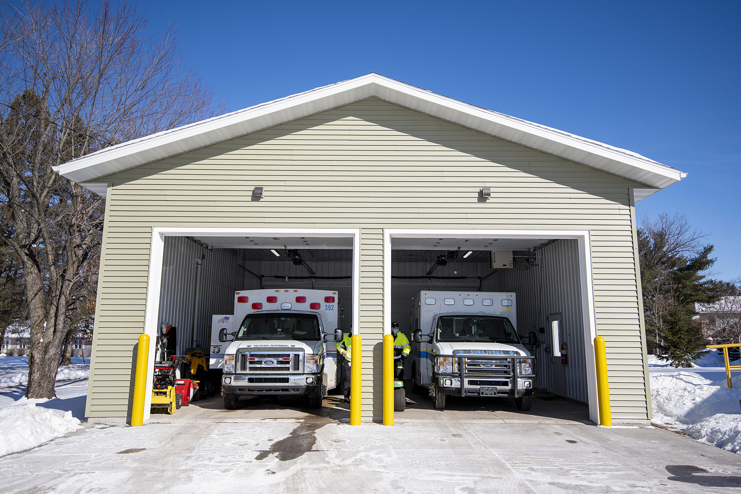 Two ambulances are parked inside of a small garage with the doors rolled up.