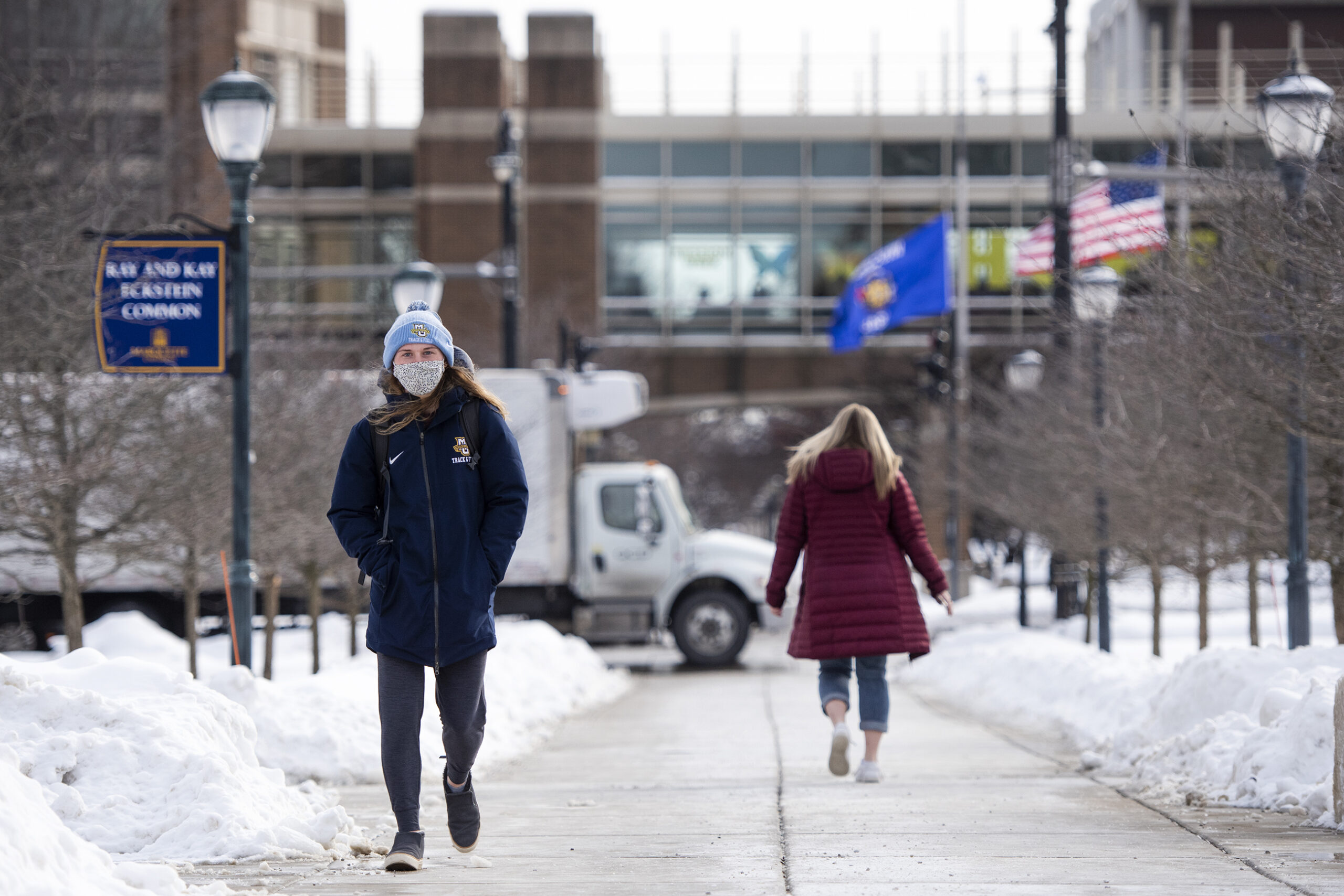 A girl wears a Marquette hat while walking in front of an elevated walkway on the campus. Snow is on the ground.