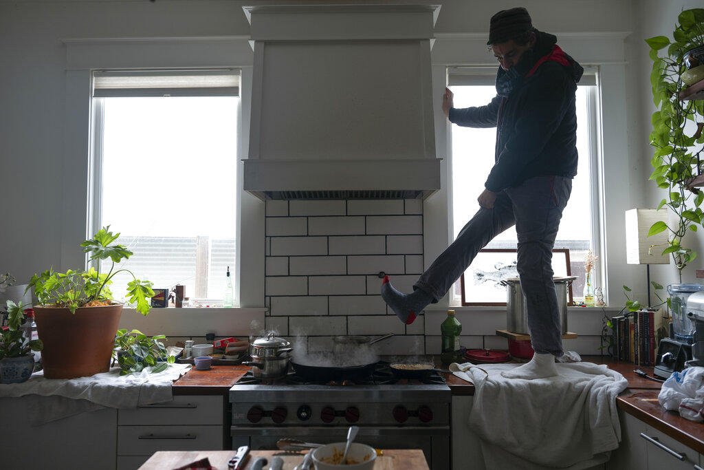 A man stands on his kitchen counter wearing many layers and hovering one boot-covered foot over a steamy stove.
